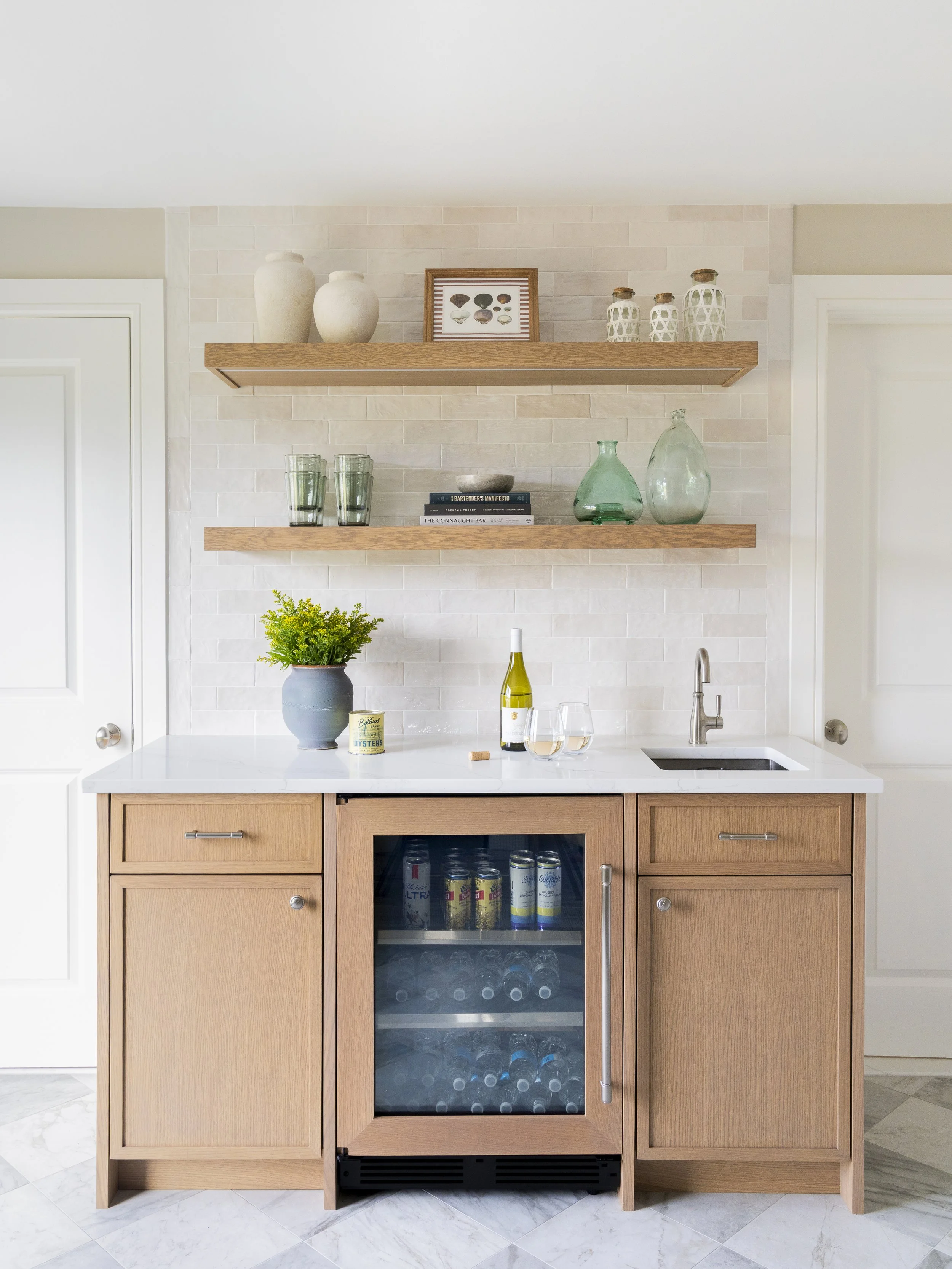 Custom mini bar with pale wood cabinetry, open shelving and beverage refrigerator designed by Blakely Interior Design.
