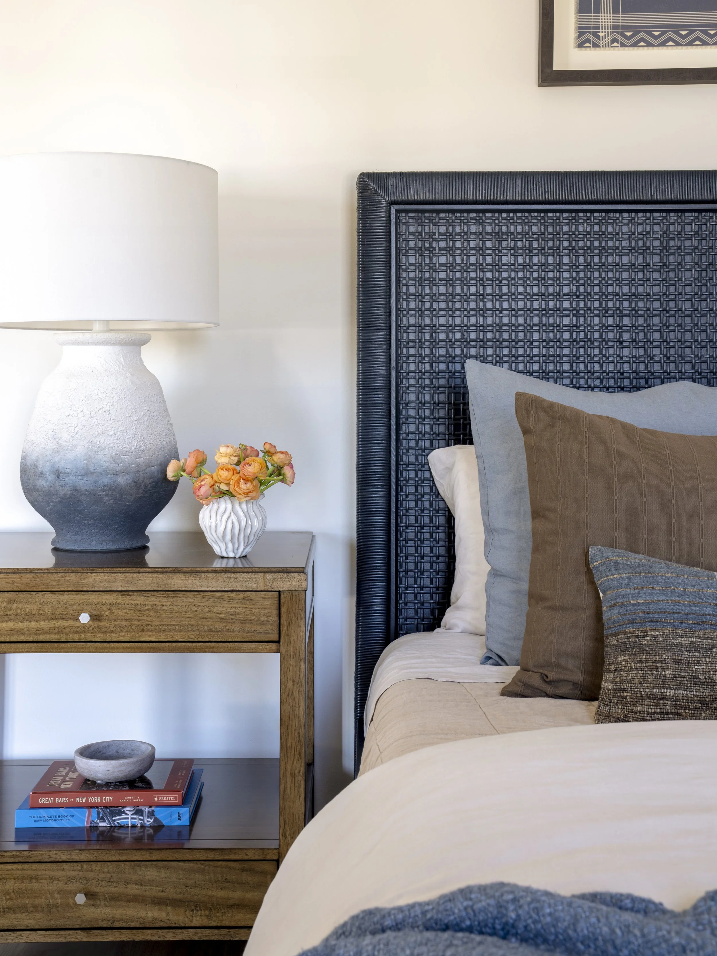 Coastal bedroom detail with woven navy headboard, wood nightstand and layered neutral bedding.