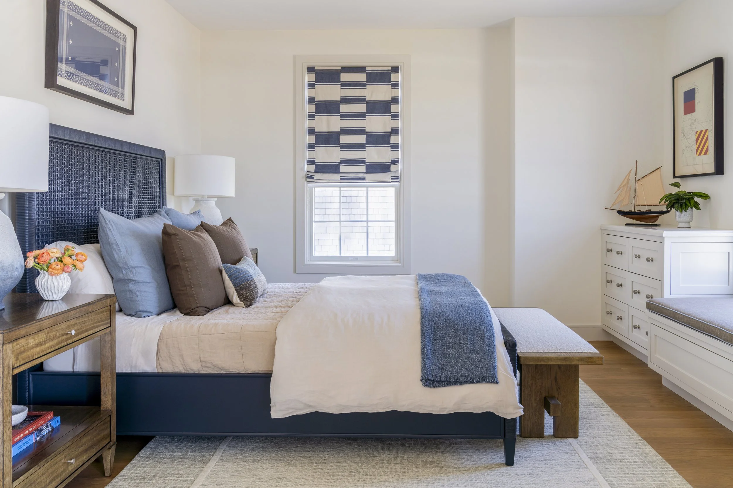Blue coastal bedroom with layered bedding, natural wood furnishings and patterned Roman shade in Narragansett, RI.