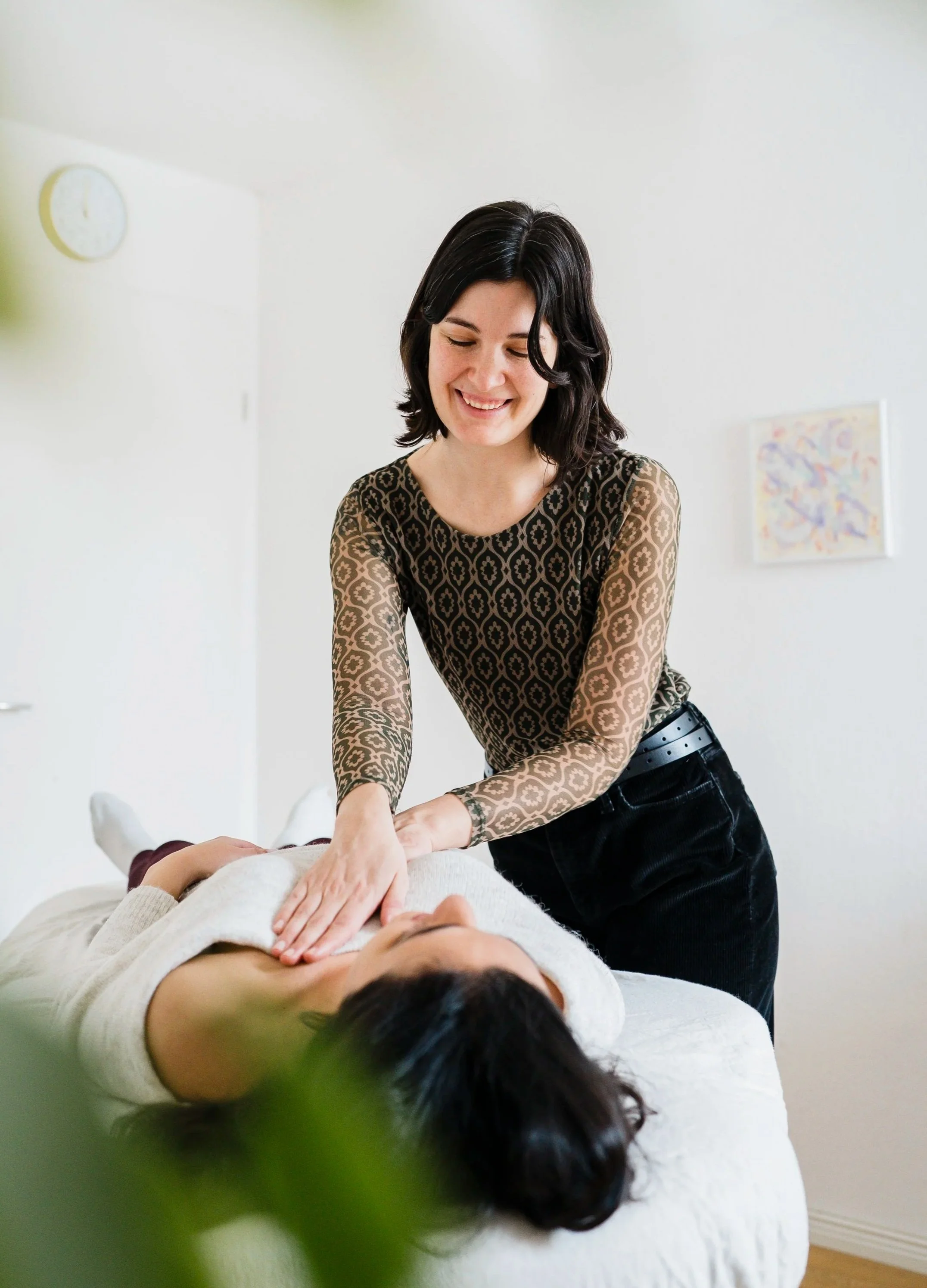 A woman giving a Pantarei Approach Bodywork session to another woman lying on a massage table, smiling in a bright, minimalist room.