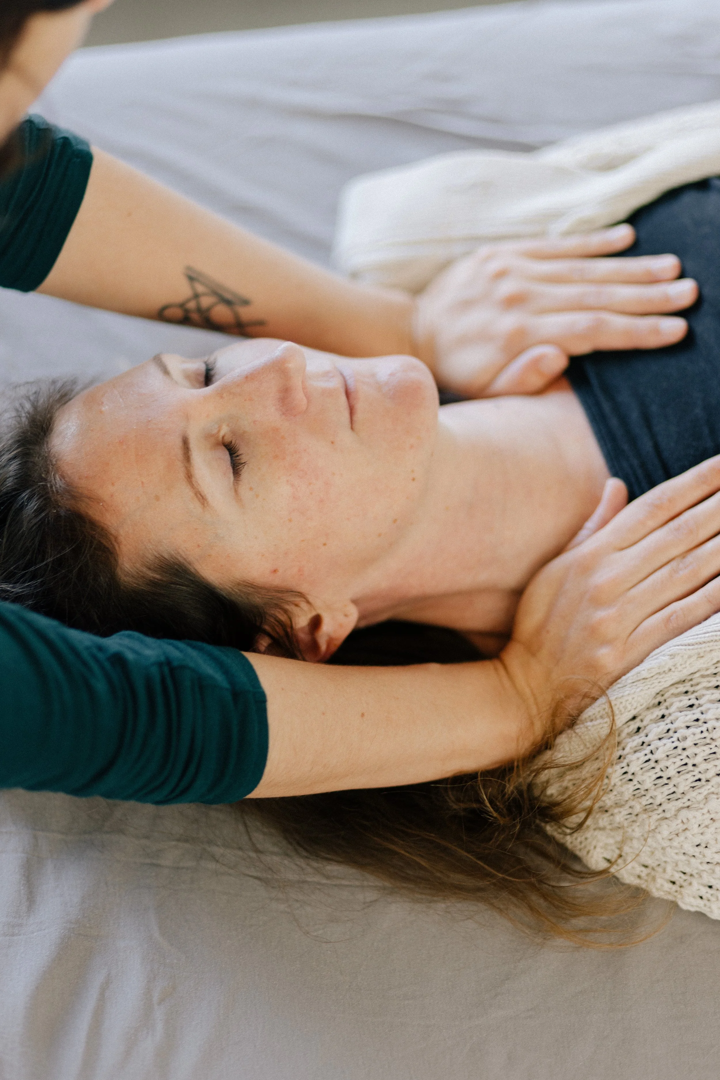 A woman with closed eyes lying down receiving a massage, with another person's hands on her chest and head.