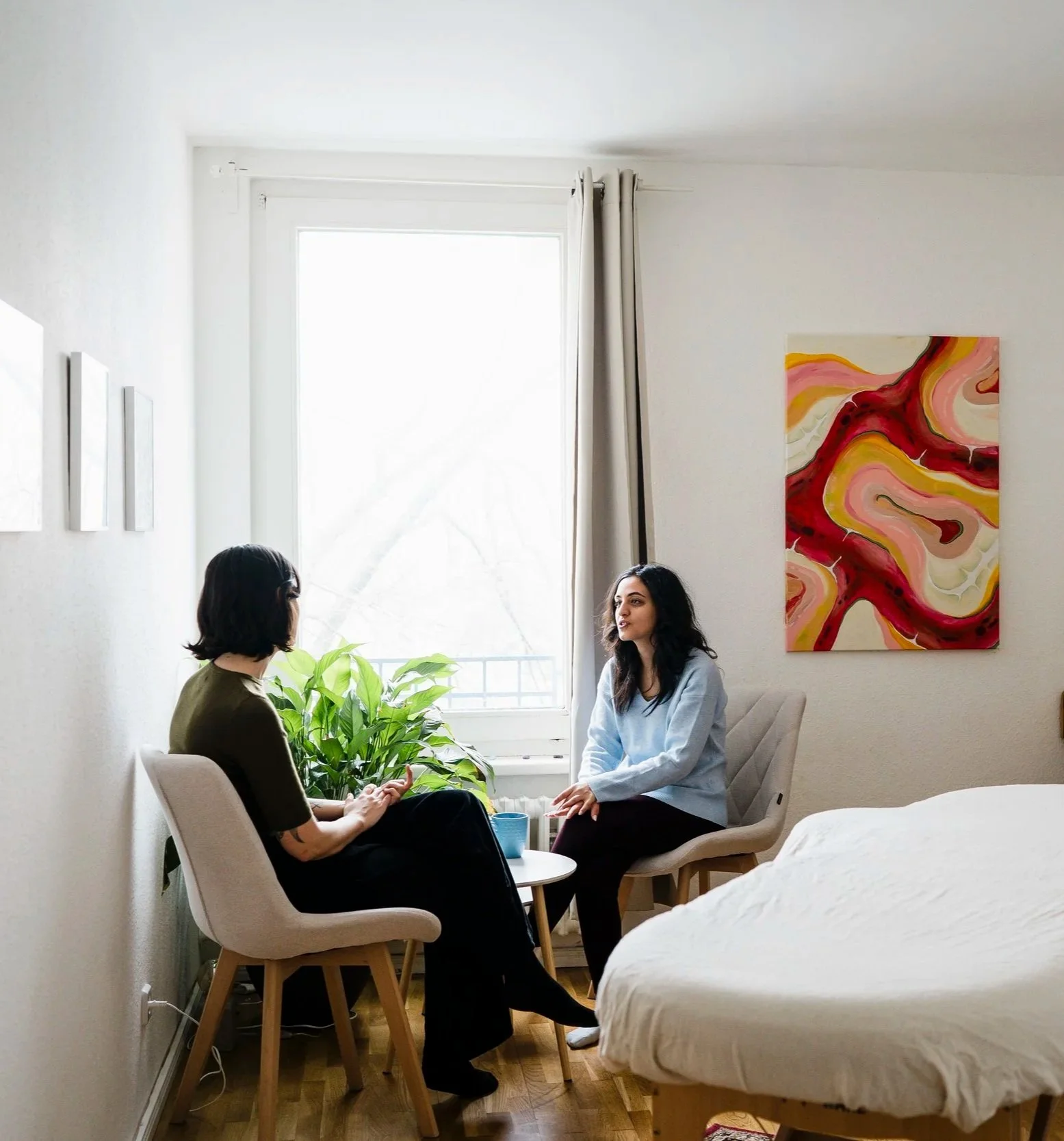 Two women having a conversation at the start of a Pantarei Approach Bodywork session. They are seated facing each other, with a potted plant and abstract painting on the wall.