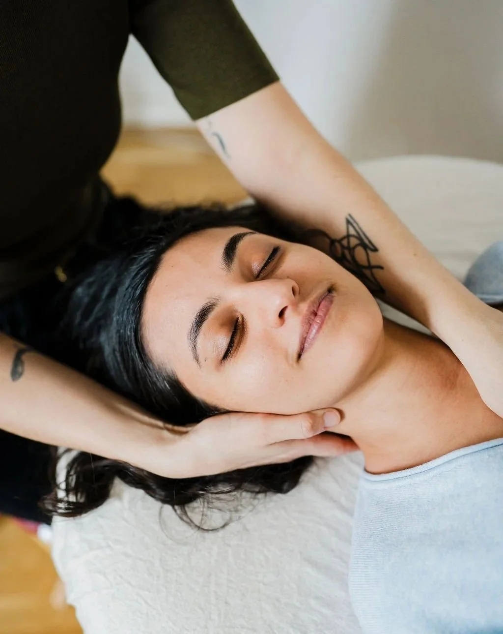 A woman with dark hair and receives a Pantarei Approach bodywork session, lying on a bed with her eyes closed and a peaceful expression.