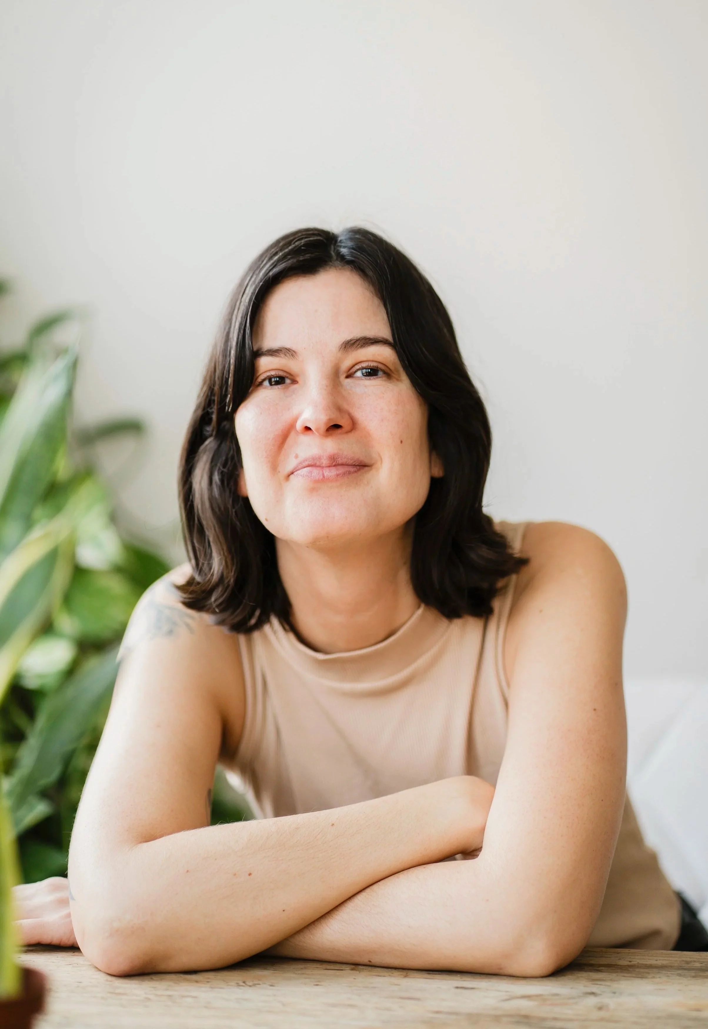 A woman with short dark hair and light skin sitting at a wooden table, smiling slightly at the camera, with a green plant in the background.