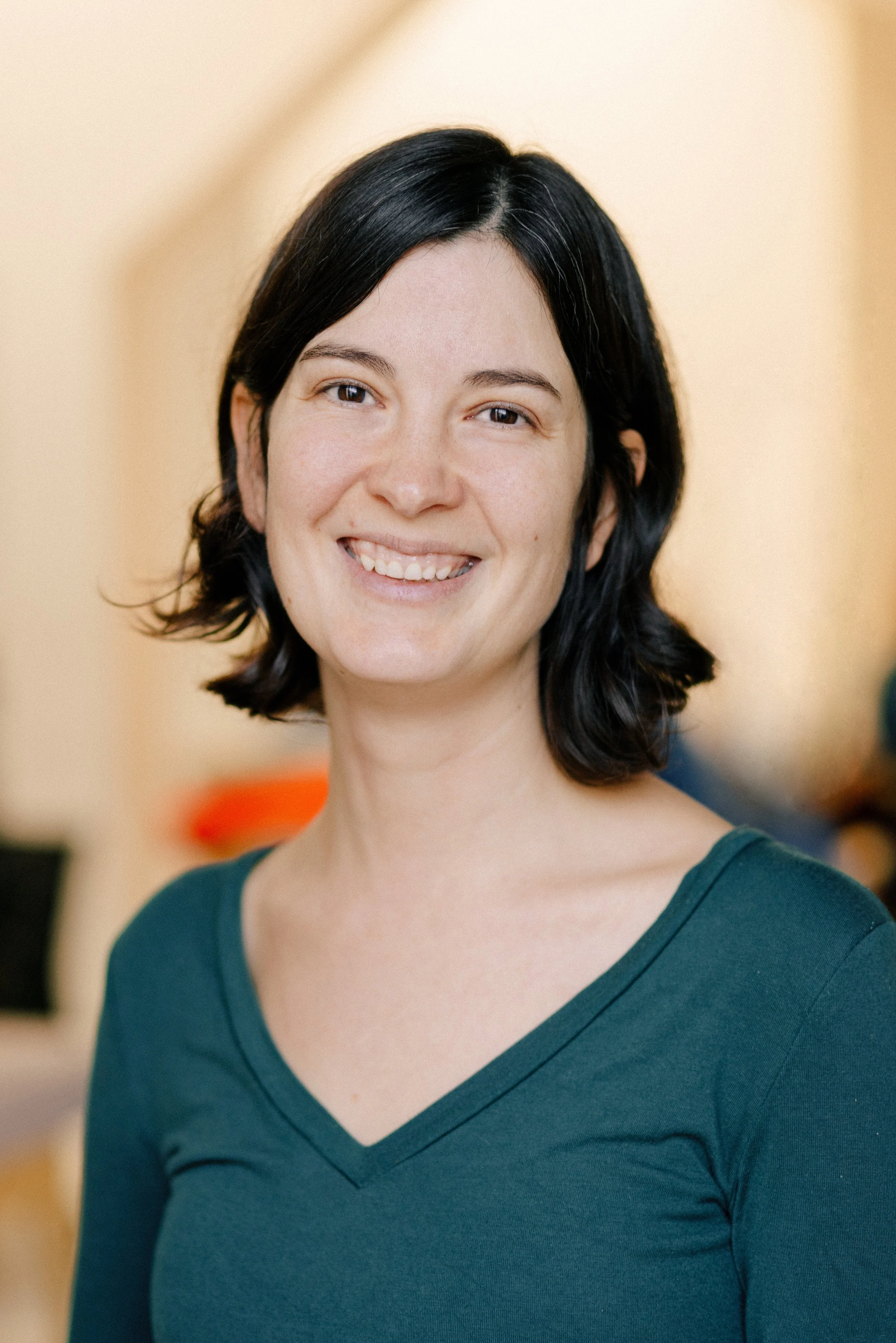 A woman with black hair smiling, wearing a teal V-neck shirt, in an indoor setting with warm-colored walls.