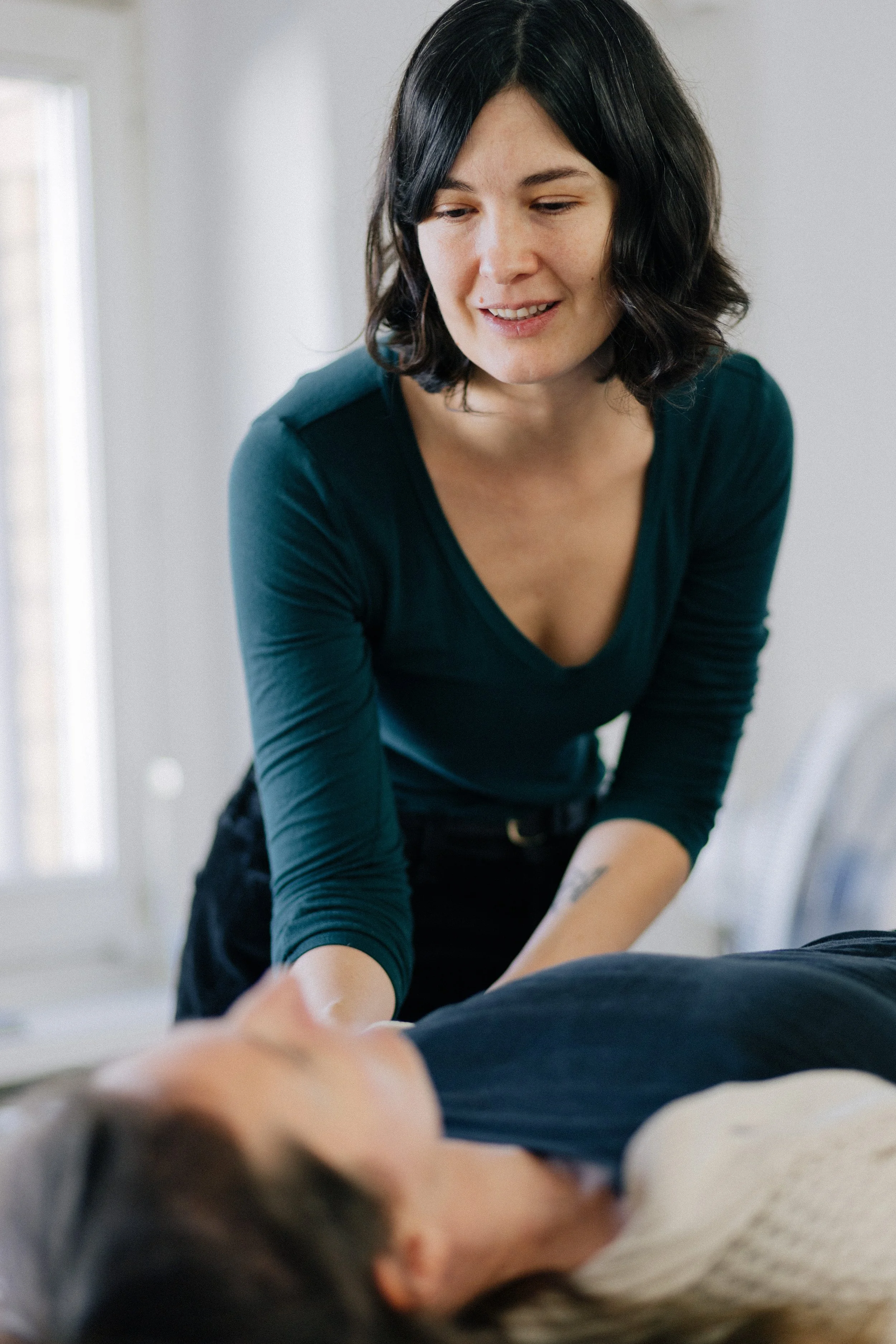A woman with dark hair smiling while giving a massage to a person lying on a bed or table.
