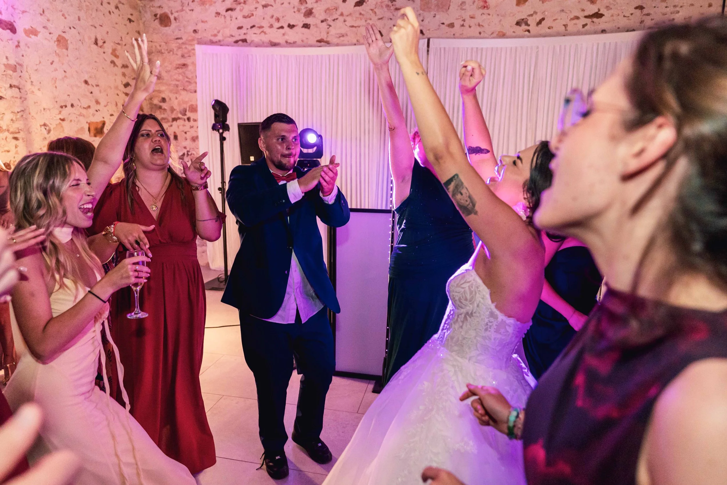 Groupe de personnes dansant et s'amusant lors d'une fête ou d'un mariage, avec un fond de murs en pierre et une scène lumineuse.