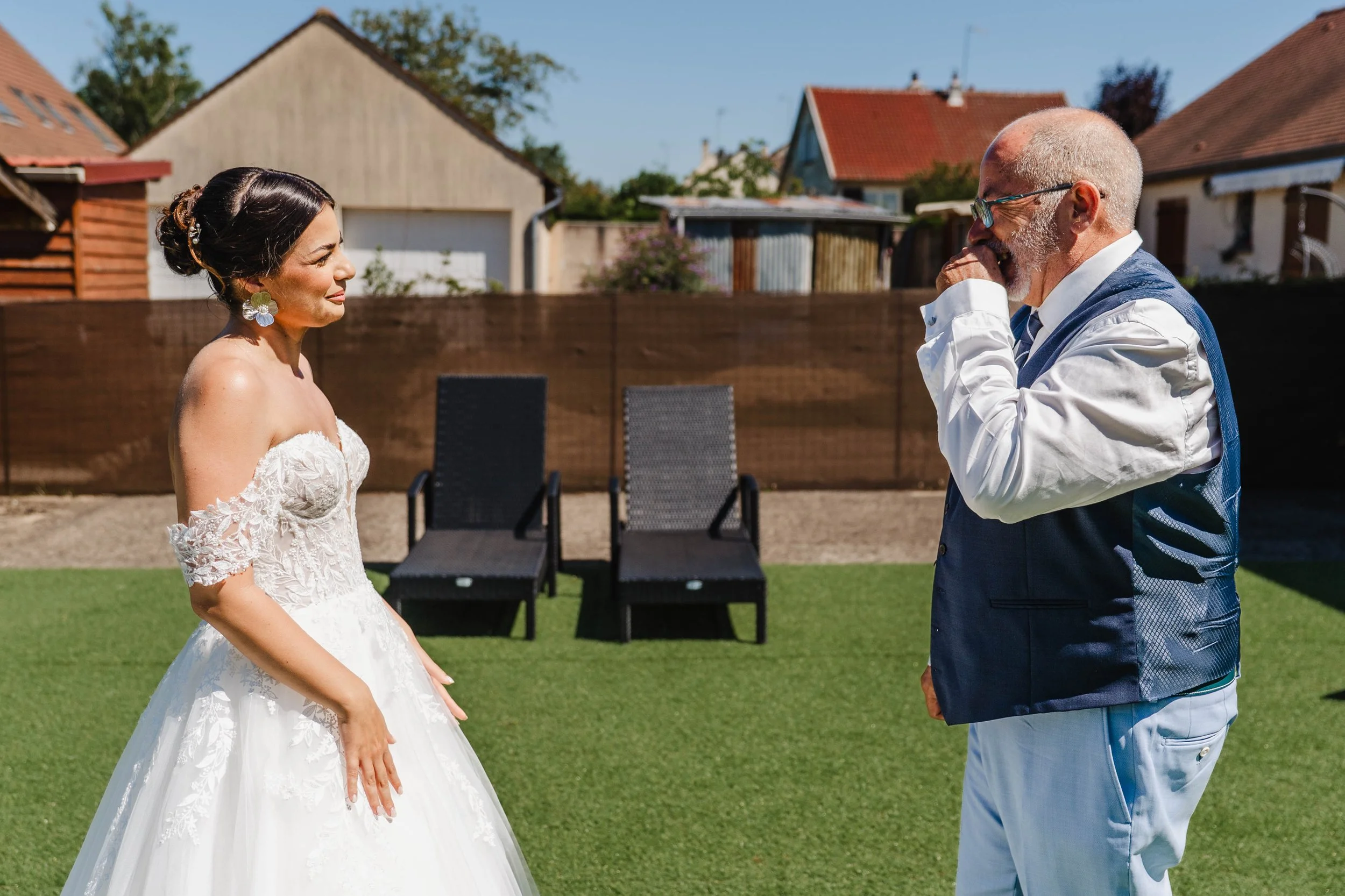 Une femme en robe de mariage sourit en face d'un homme habillé de façon élégante dans un jardin sous un ciel clair.
