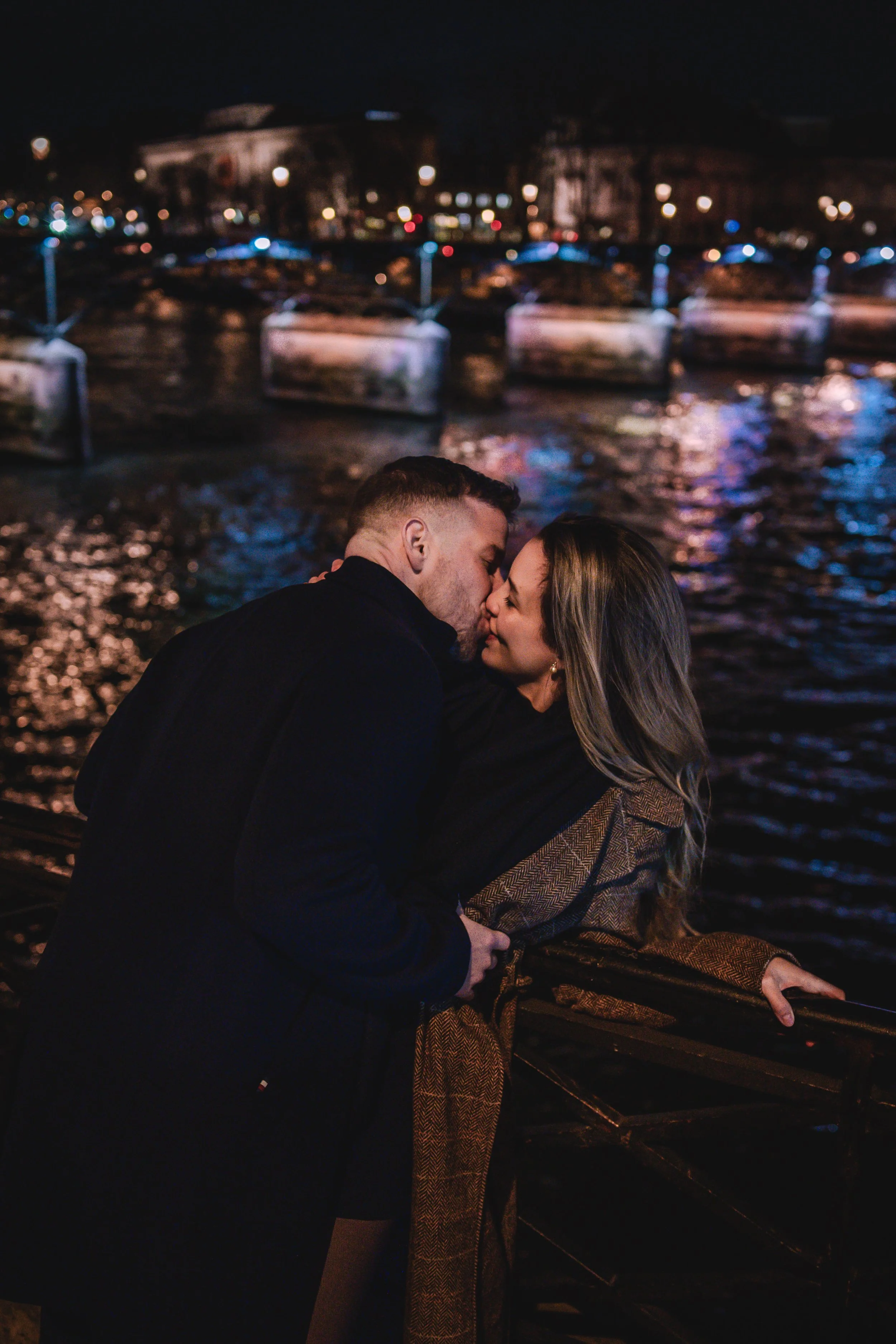 Un couple s'embrasse près d'une rivière la nuit, avec un pont éclairé en fond.