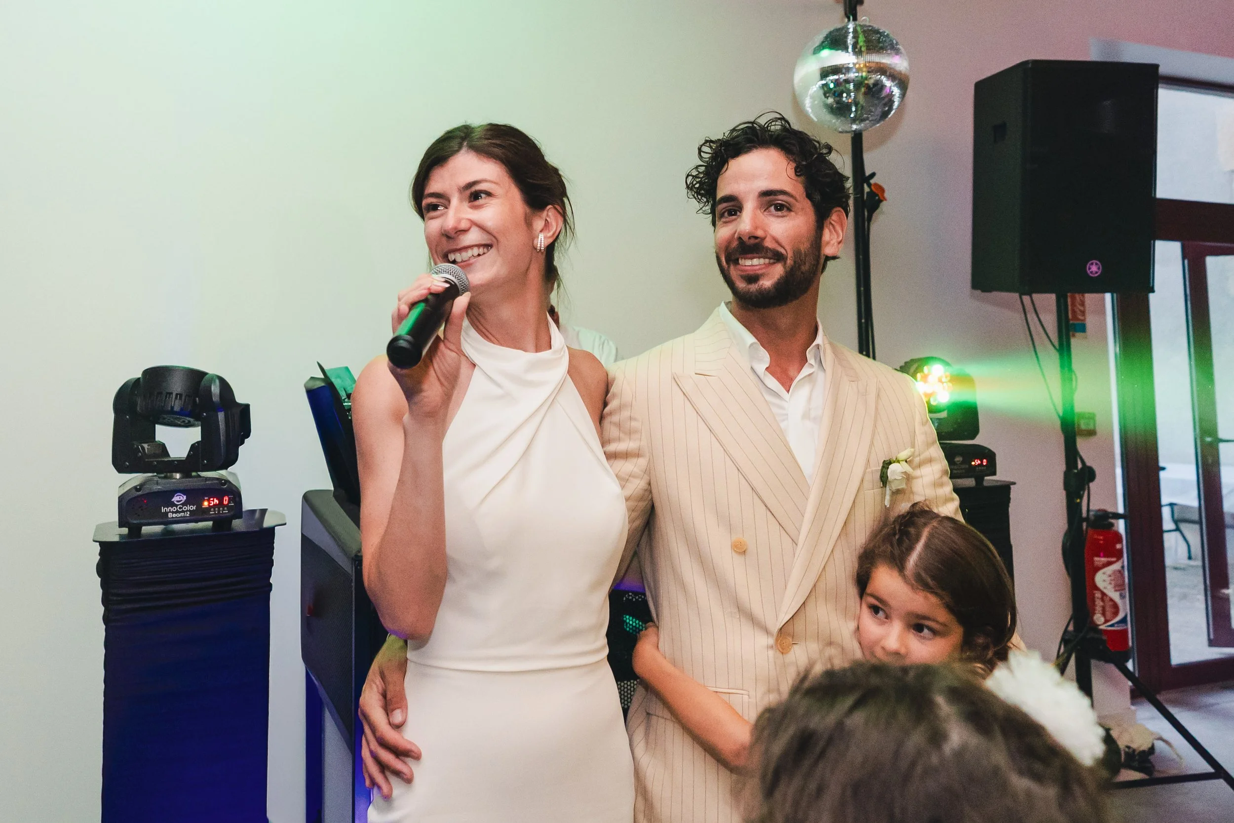 Un couple habillé élégamment lors d'un mariage, la femme avec une robe blanche et le homme avec un costume beige, entourés de personnes dont une jeune fille avec un bouquet, dans une salle avec des lumières et un système de sonorisation.