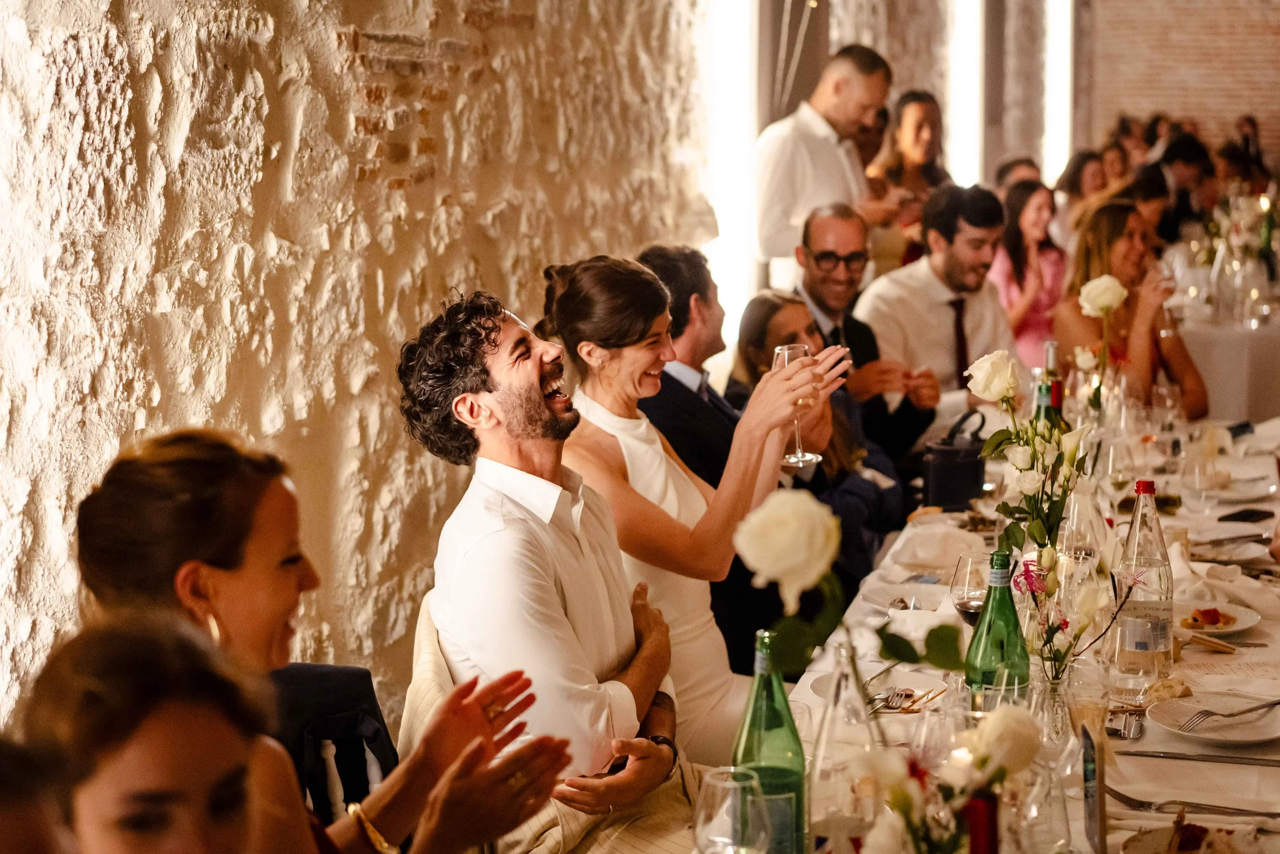 Groupe de personnes assises à une longue table lors d'une célébration, certains riant et souriant, ambiance chaleureuse avec décor floral et assiettes.