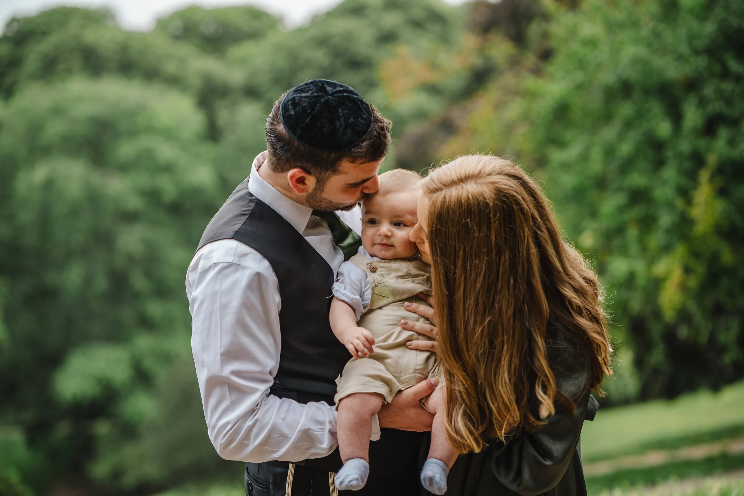 Un couple, un homme portant une kippa noire et une femme aux cheveux longs, tiennent un bébé dans le parc des buttes chaumont, échangeant un baiser avec le bébé.