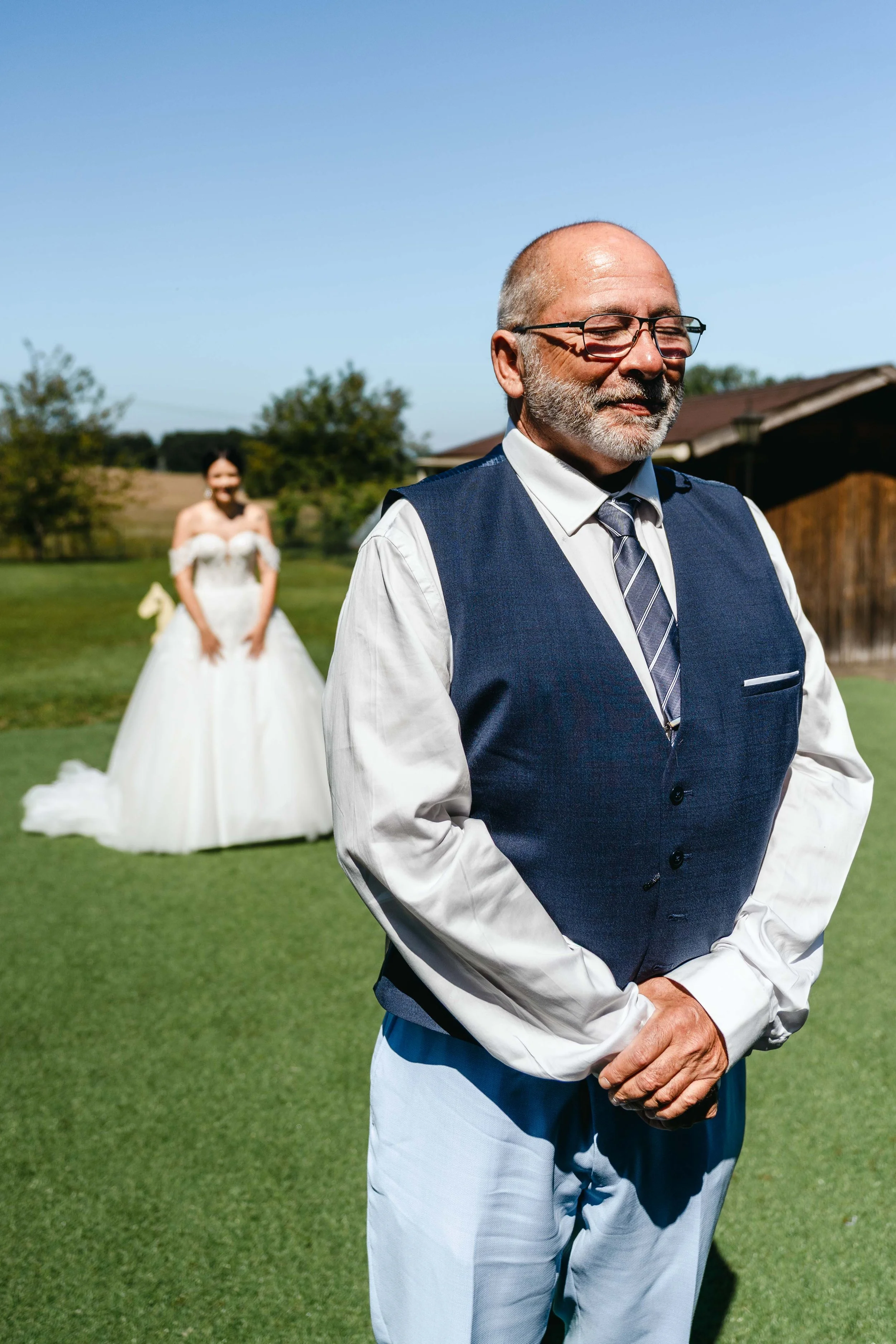 Un homme âgé, vêtu d'une chemise blanche, d'un gilet bleu et d'une cravate, se tient avec ses mains jointes devant lui sur une pelouse verte, avec une femme en robe de mariée en arrière-plan dans un jardin en plein air sous un ciel bleu clair.