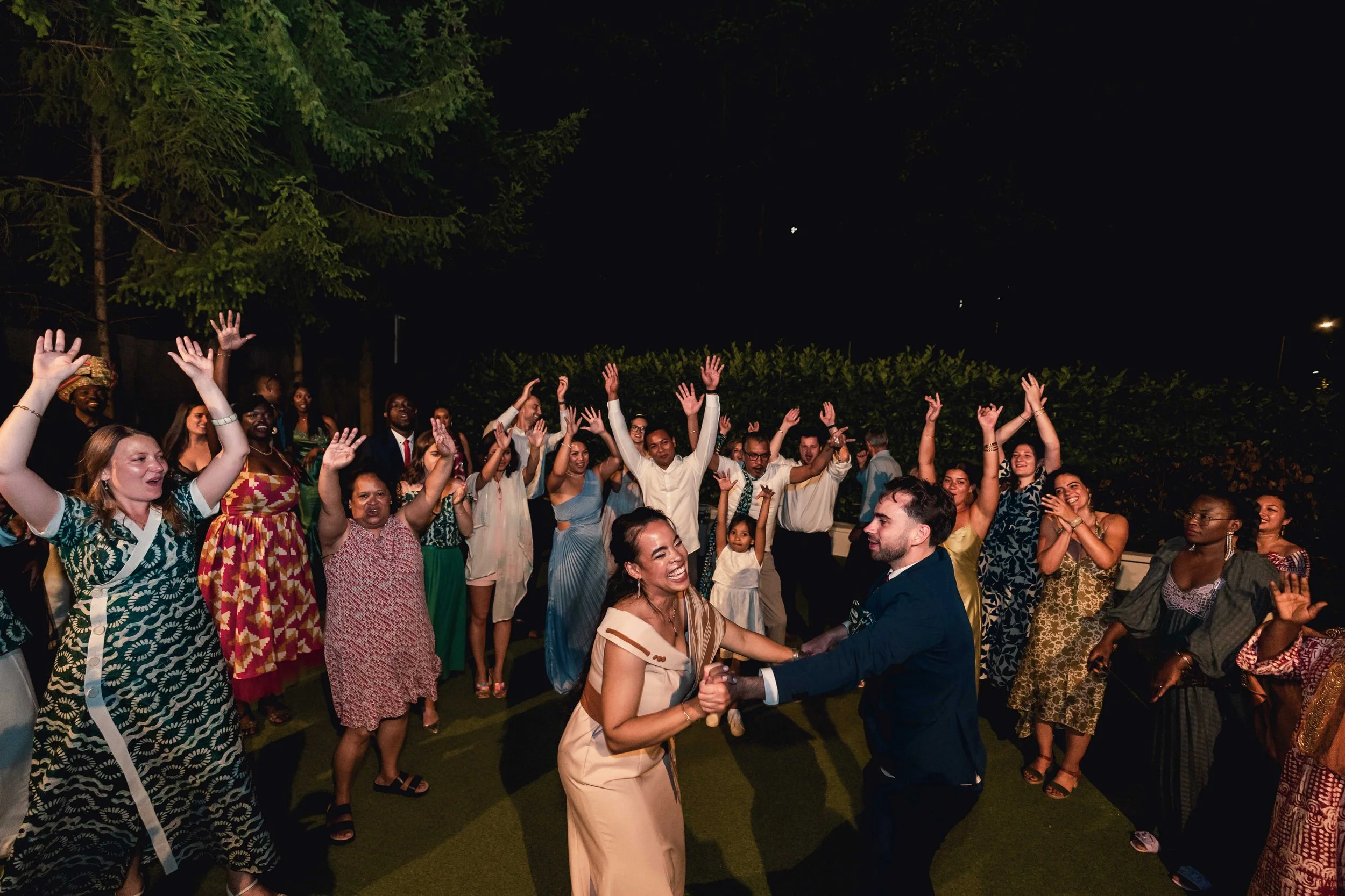 Un groupe de personnes danse et s'amuse lors d'une fête nocturne en plein air, avec deux personnes au centre qui dansent ensemble, entourées d'autres invités qui lèvent les bras et sourient.