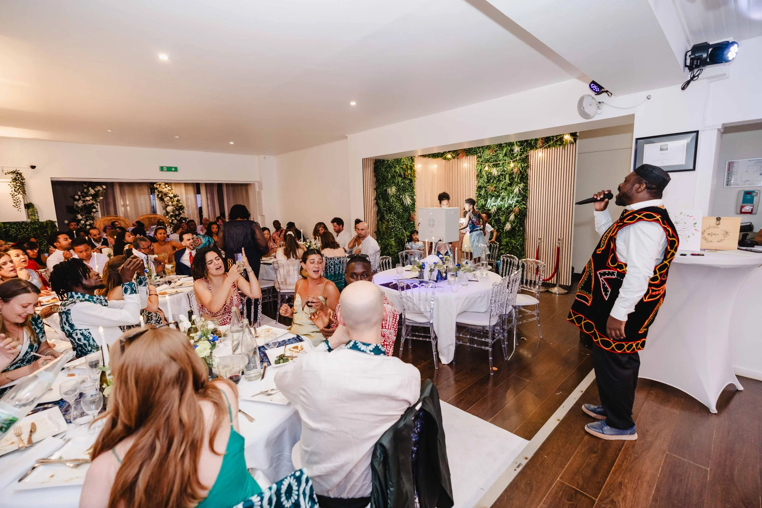 Un groupe de personnes assises autour de tables lors d'un événement, avec un homme chantant avec un microphone dans un coin, décorations florales et verdure en arrière-plan.