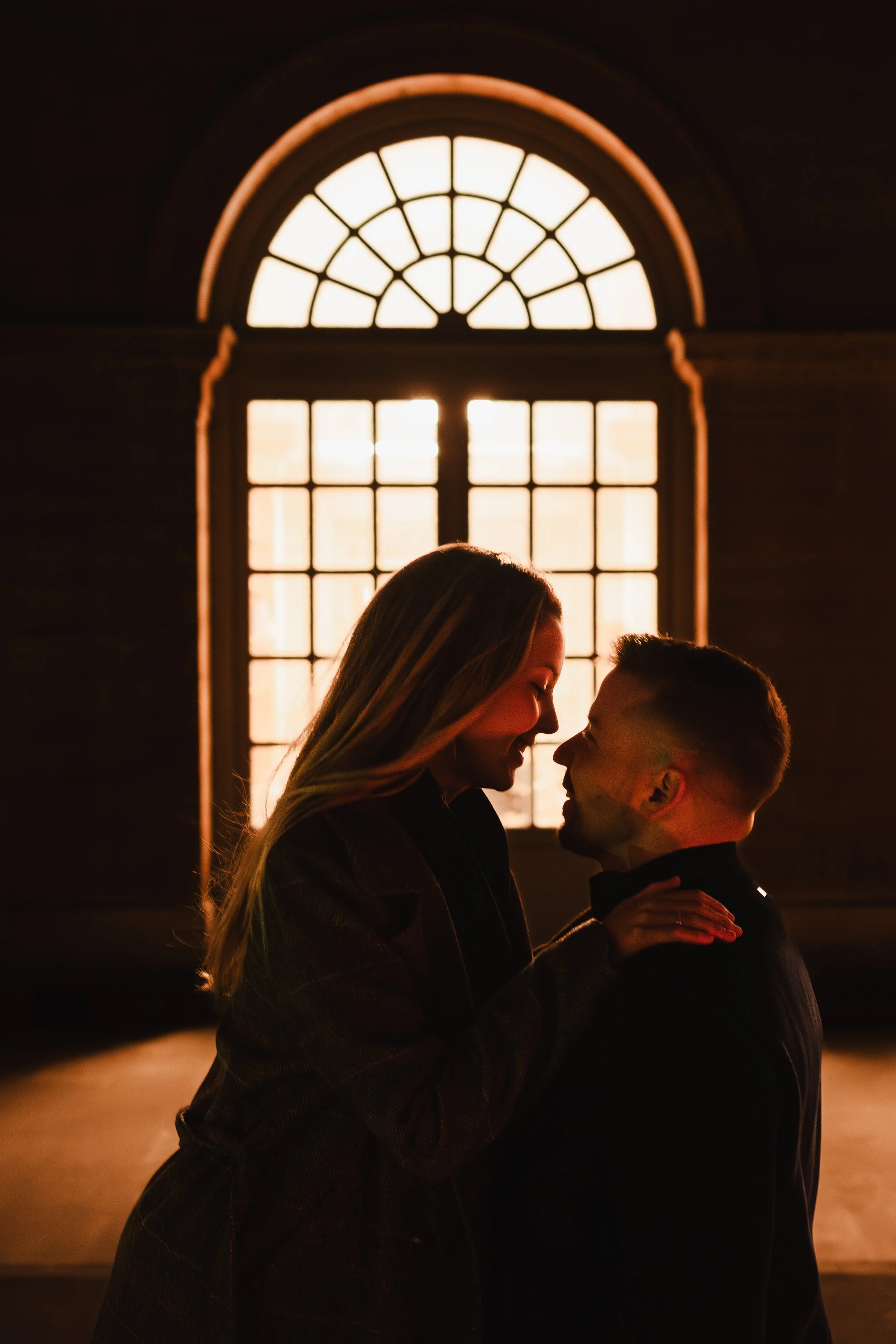 Un couple s'embrasse près d'une grande fenêtre lumineuse, devant le palais Royal, créant un contraste chaud et romantique.