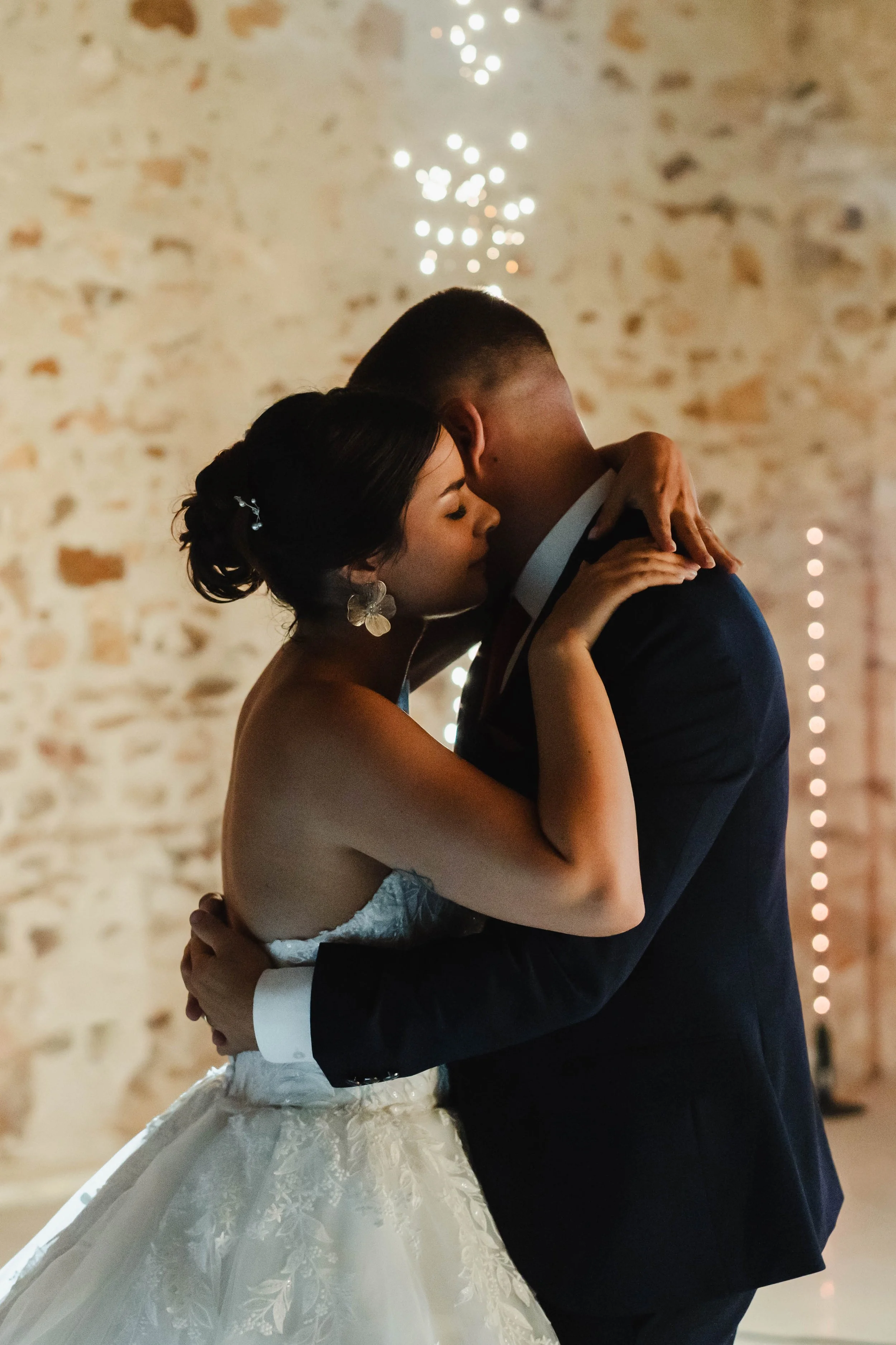 Un couple danse lors de leur mariage dans une salle avec un mur en pierre et des lumières décoratives.