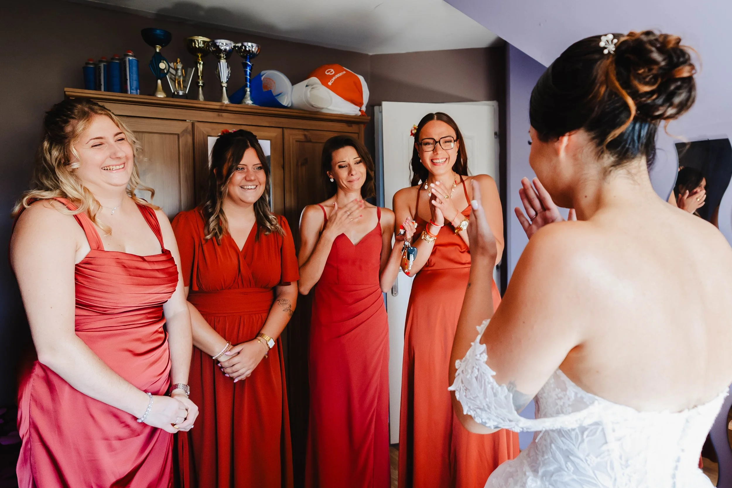 Fiançailles dans une pièce avec cinq femmes habillées en rouge, souriantes et émues, voyant la mariée en robe de mariée.