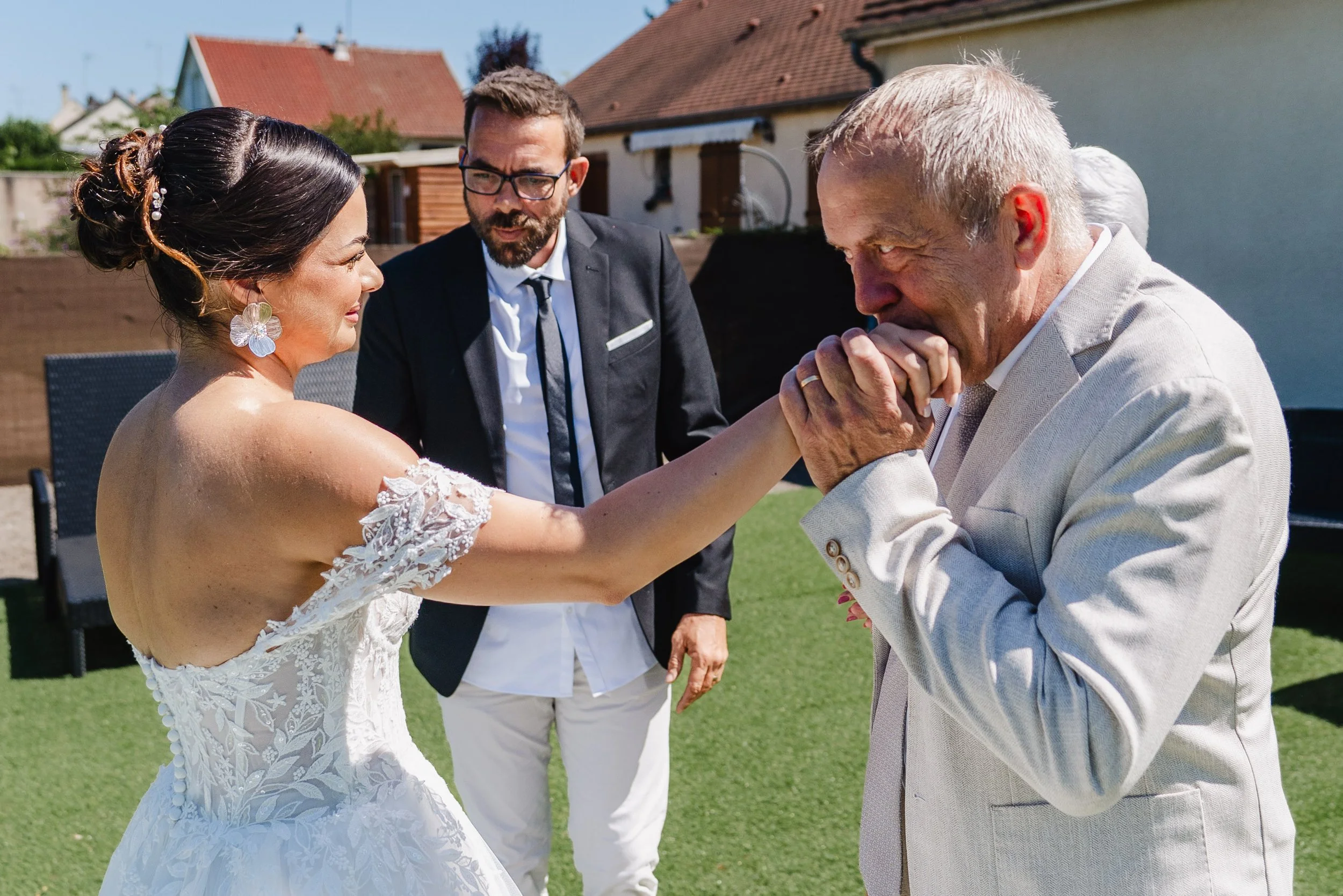 Une femme en robe de mariage embrasse un homme âgé lors d'une cérémonie en plein air, avec un homme en costume noir observant la scène.