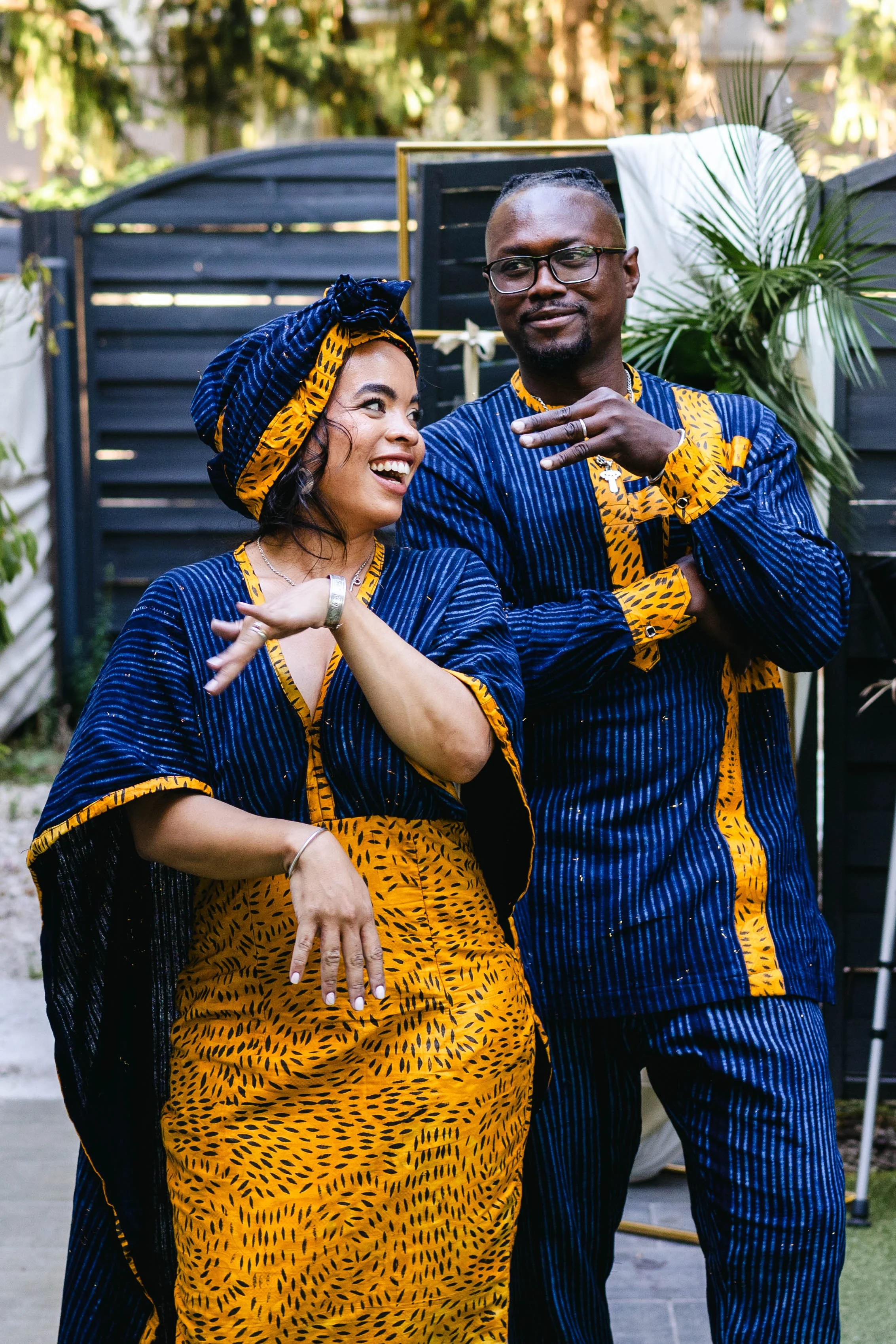 Un couple habillé en vêtements traditionnels colorés avec des motifs géométriques, l'homme porte des lunettes et des bagues, ils sourient et semblent danser ou célébrer dans un jardin avec des plantes vertes.
