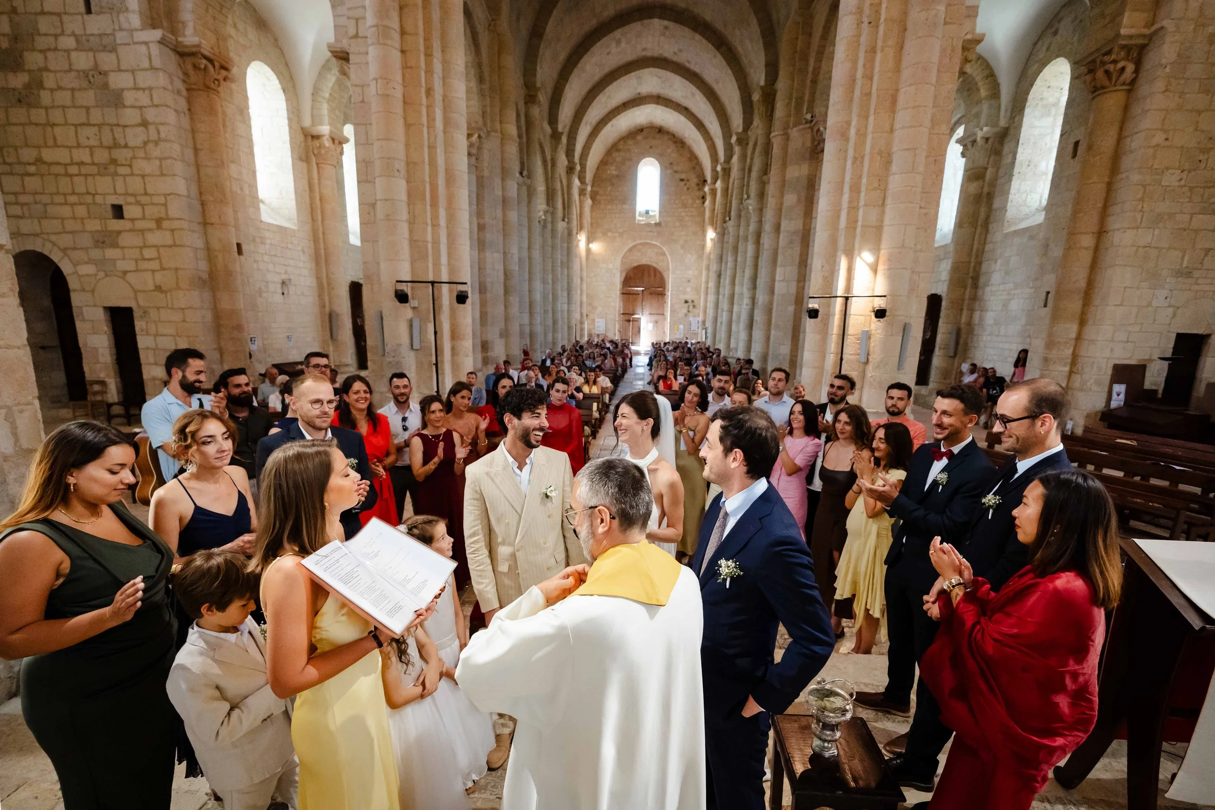 Cérémonie de mariage dans l'église en pierre de Moirax , avec un couple devant le prêtre et de nombreux invités regardant et applaudissant.