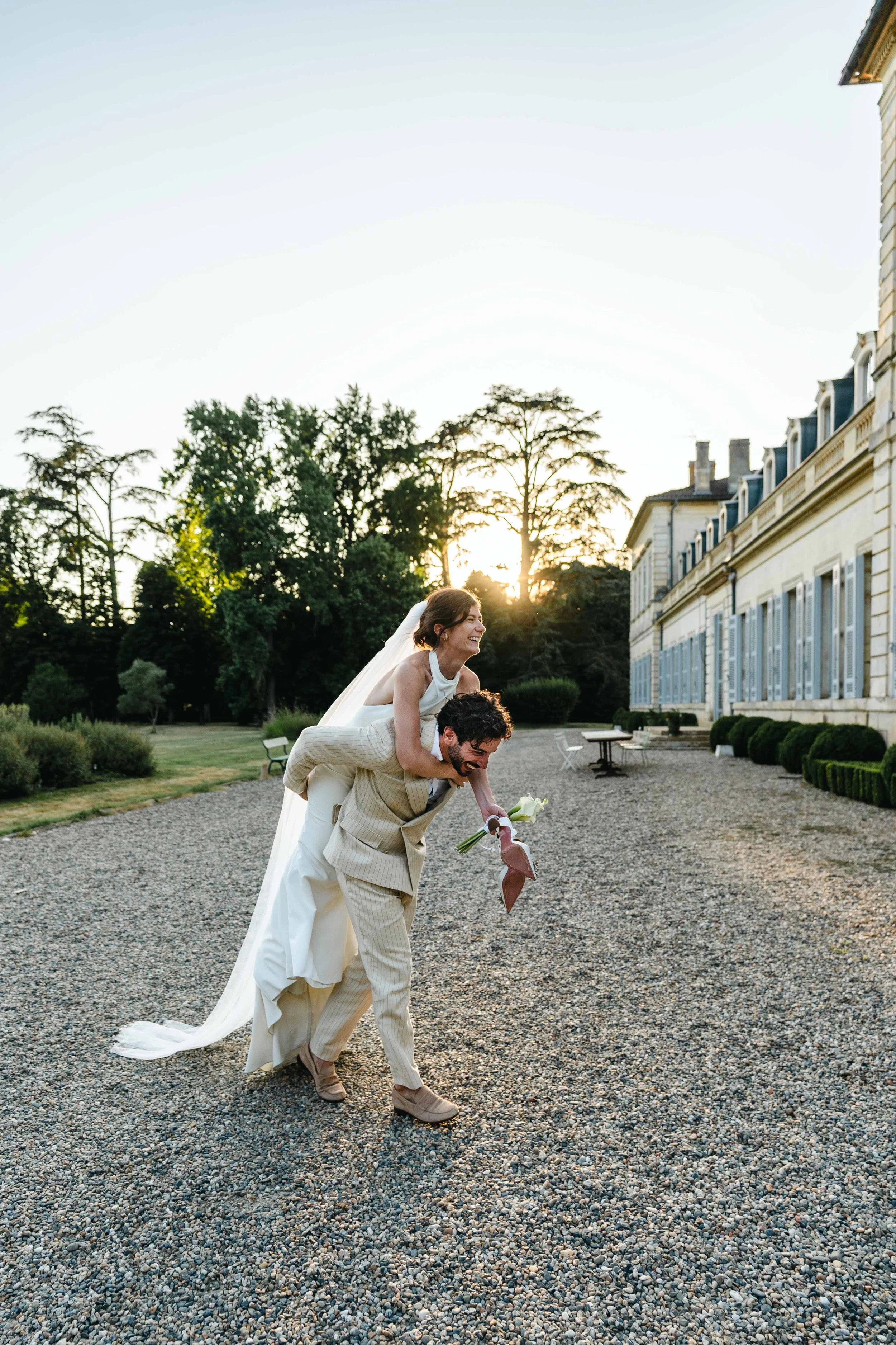 Un couple de mariés riant, la femme étant portée sur le dos de l'homme, en plein air au chateau Saint Denis au coucher du soleil.