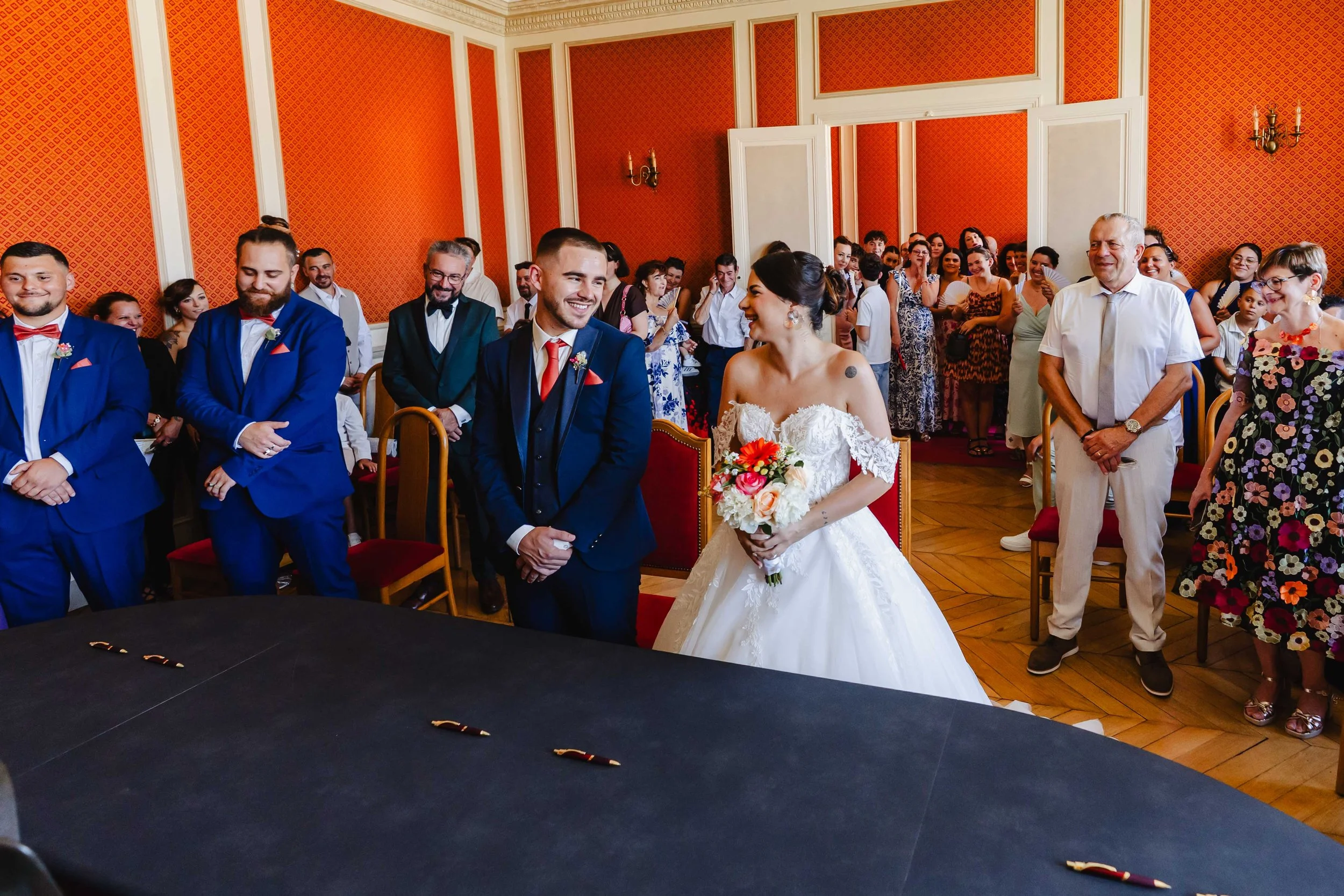 Un couple de mariés lors d'une cérémonie de mariage entouré d'invités dans une salle élégante avec des murs orange.