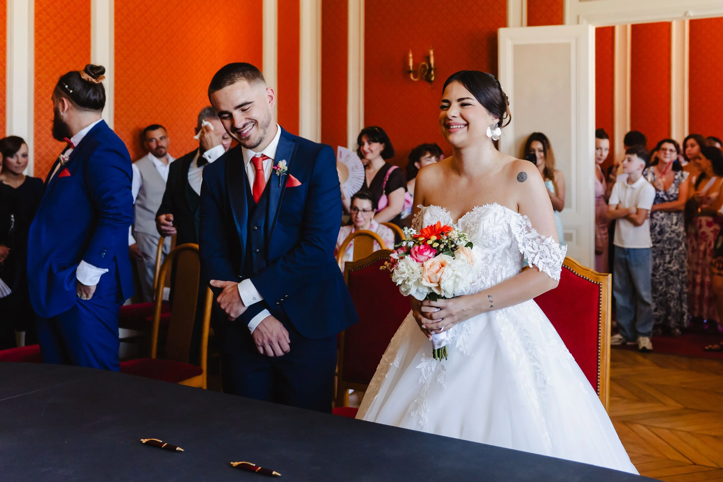 Un couple de mariés lors de leur cérémonie, la mariée en robe blanche avec un bouquet de fleurs, le marié en costume bleu, entourés de leurs proches dans une salle ornée, avec des invités en arrière-plan.