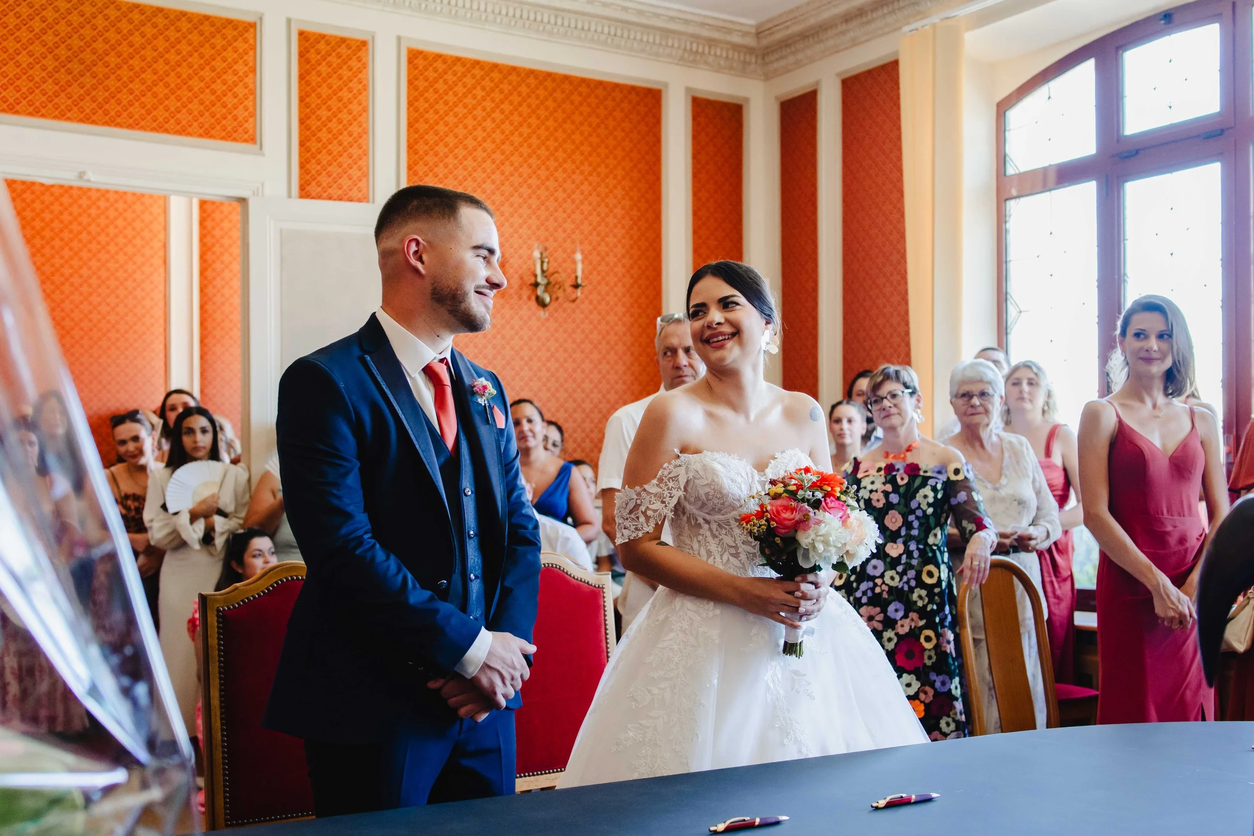 Un mariage avec le couple au centre : l'homme en costume bleu et la femme en robe de mariée blanche tenant un bouquet de fleurs, entourés de leurs invités dans une salle ornée de murs orange et de grandes fenêtres à Bagneux Sur Loing..