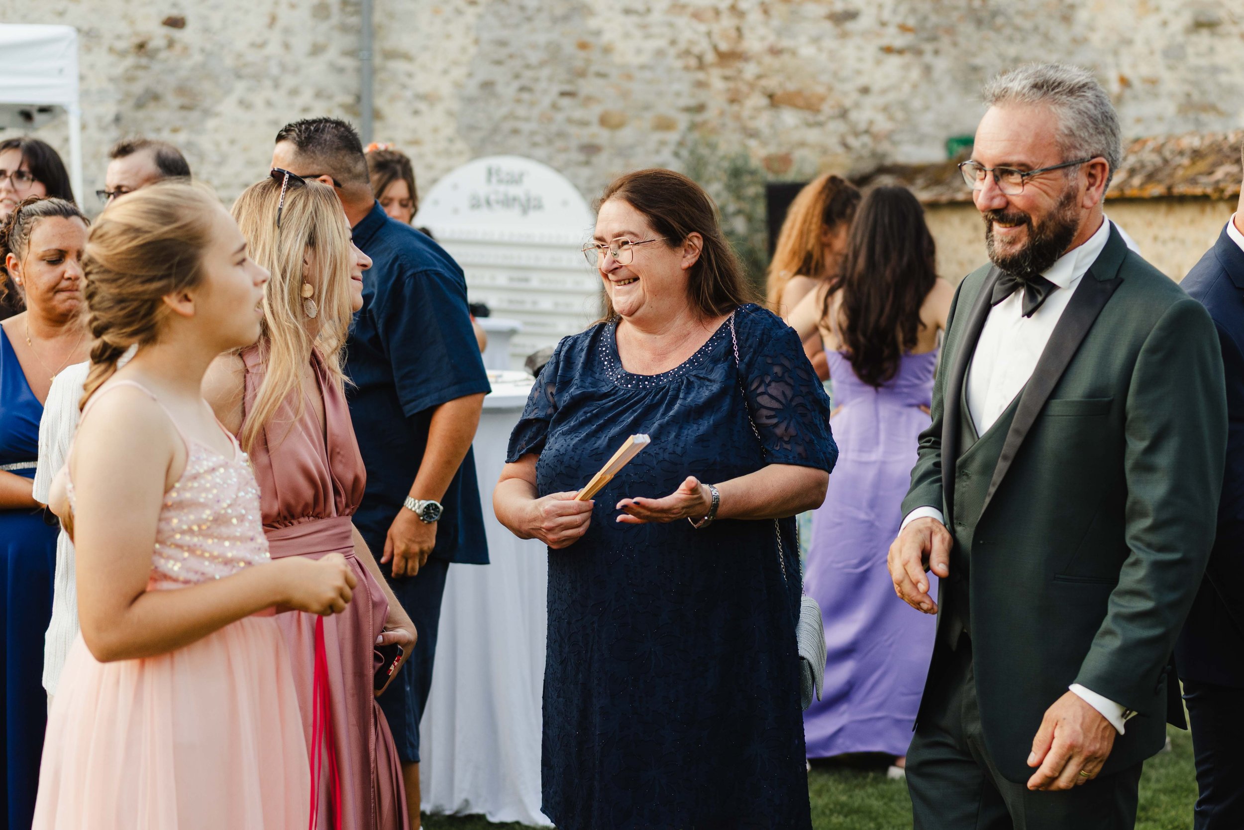 Un groupe de personnes habillées élégamment, apparemment lors d'un mariage ou d'une fête, en train de discuter et de sourire en plein air.