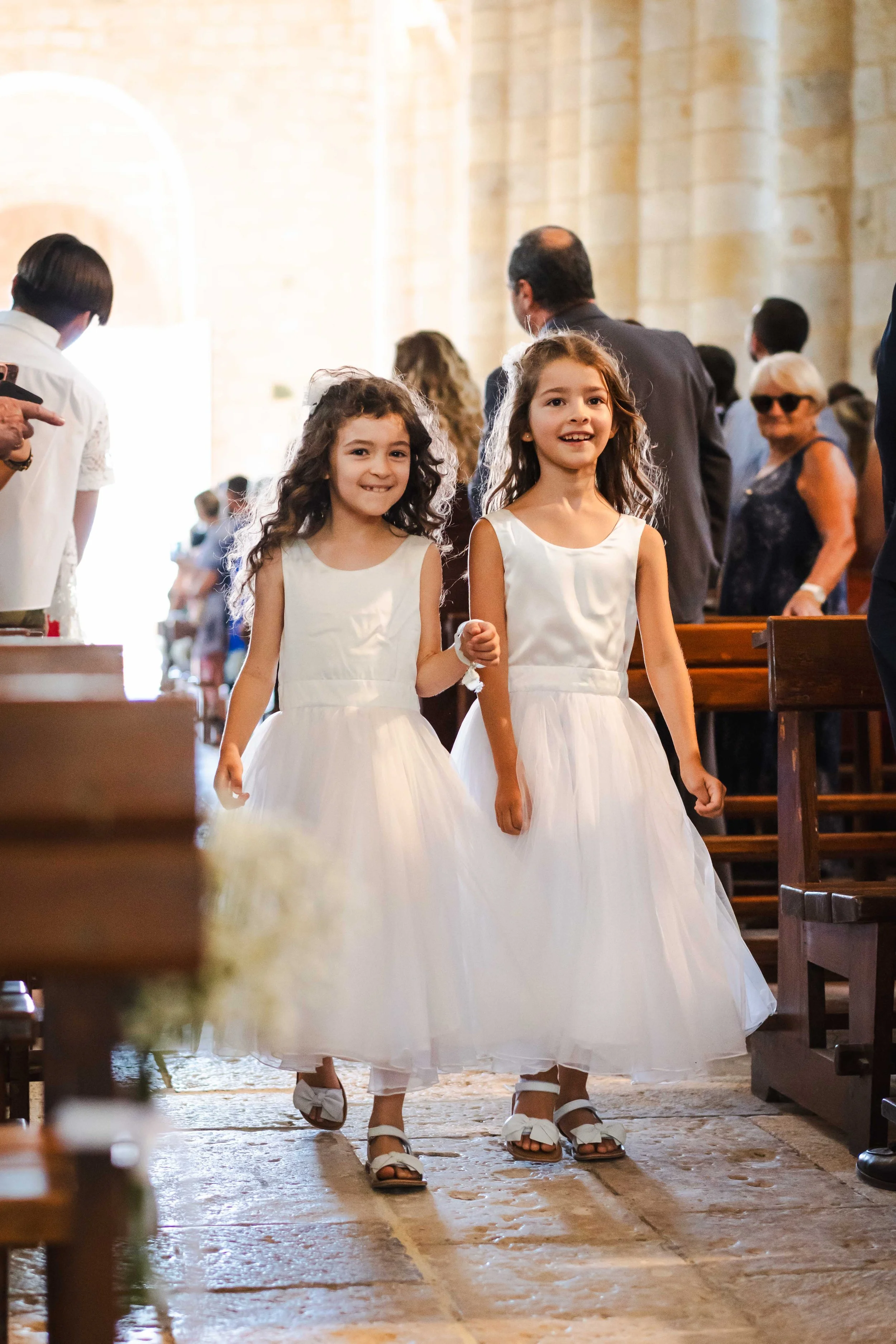 Deux jeunes filles en robes de communion blanches marchent dans une église, entourées de personnes assises et debout.