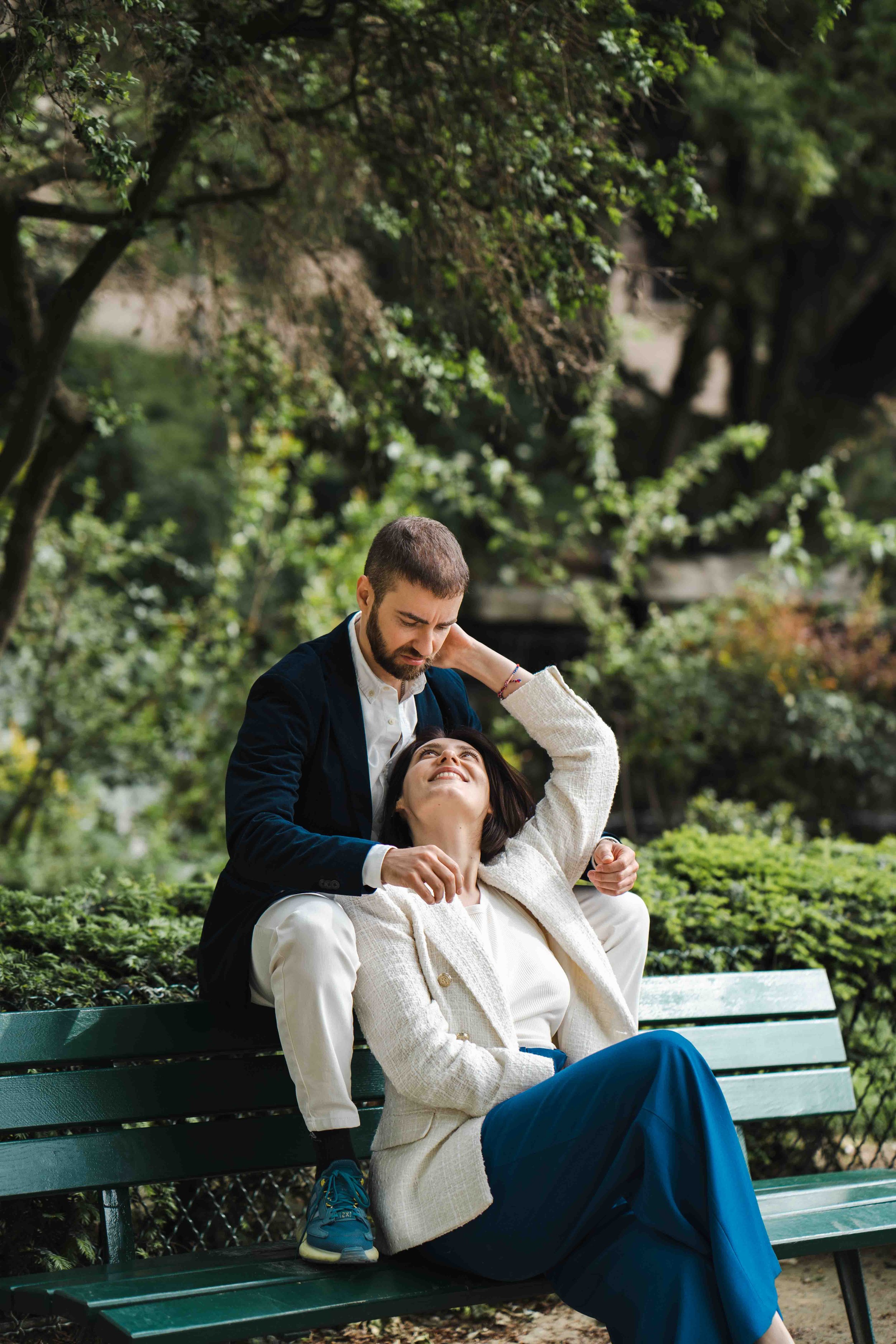 Un homme assis derrière une femme qui est assise sur un banc dans un parc, elle sourit et regarde vers le haut, lui tient la tête et la regarde avec tendresse, entourés de verdure et d'arbres.