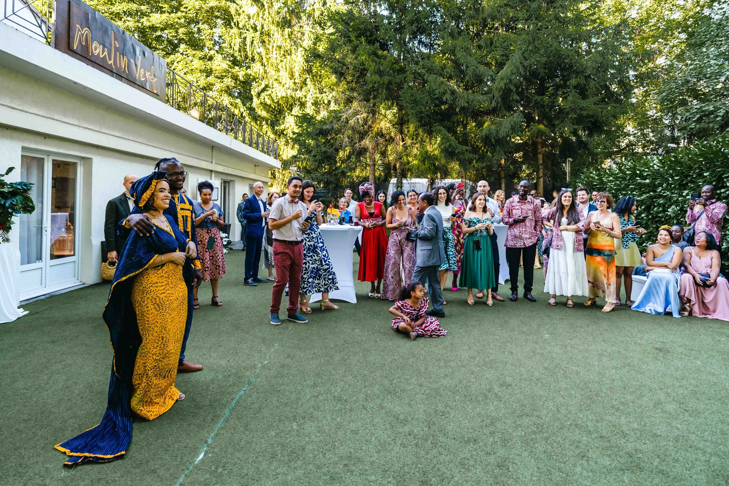Groupe de personnes à une fête en extérieur, Some de belles tenues traditionnelles et modernes, décor convivial avec arbres en arrière-plan.