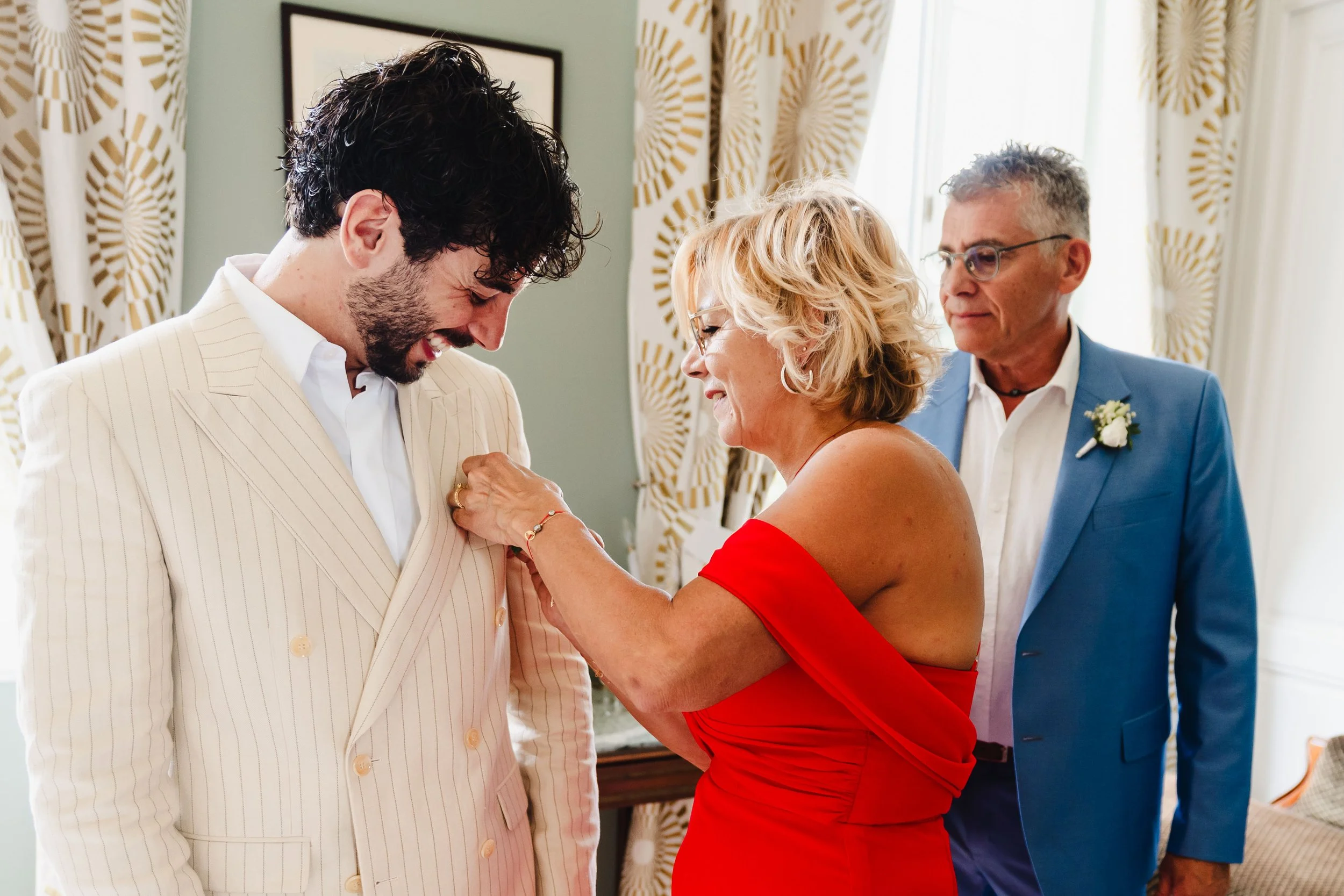 Une femme met une boutonnière à un jeune homme en costume blanc lors d'une cérémonie célébrant un mariage ou une occasion spéciale, avec un homme en veste bleue en arrière-plan dans une pièce décorée.