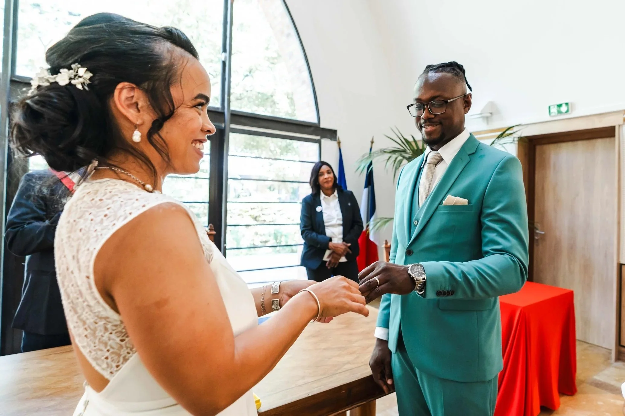 Un couple en train de se marier échange des alliances dans une cérémonie, avec une femme en costume traditionnel blanc et un homme en costume turquoise, dans une pièce lumineuse avec des fenêtres et des drapeaux derrière eux.