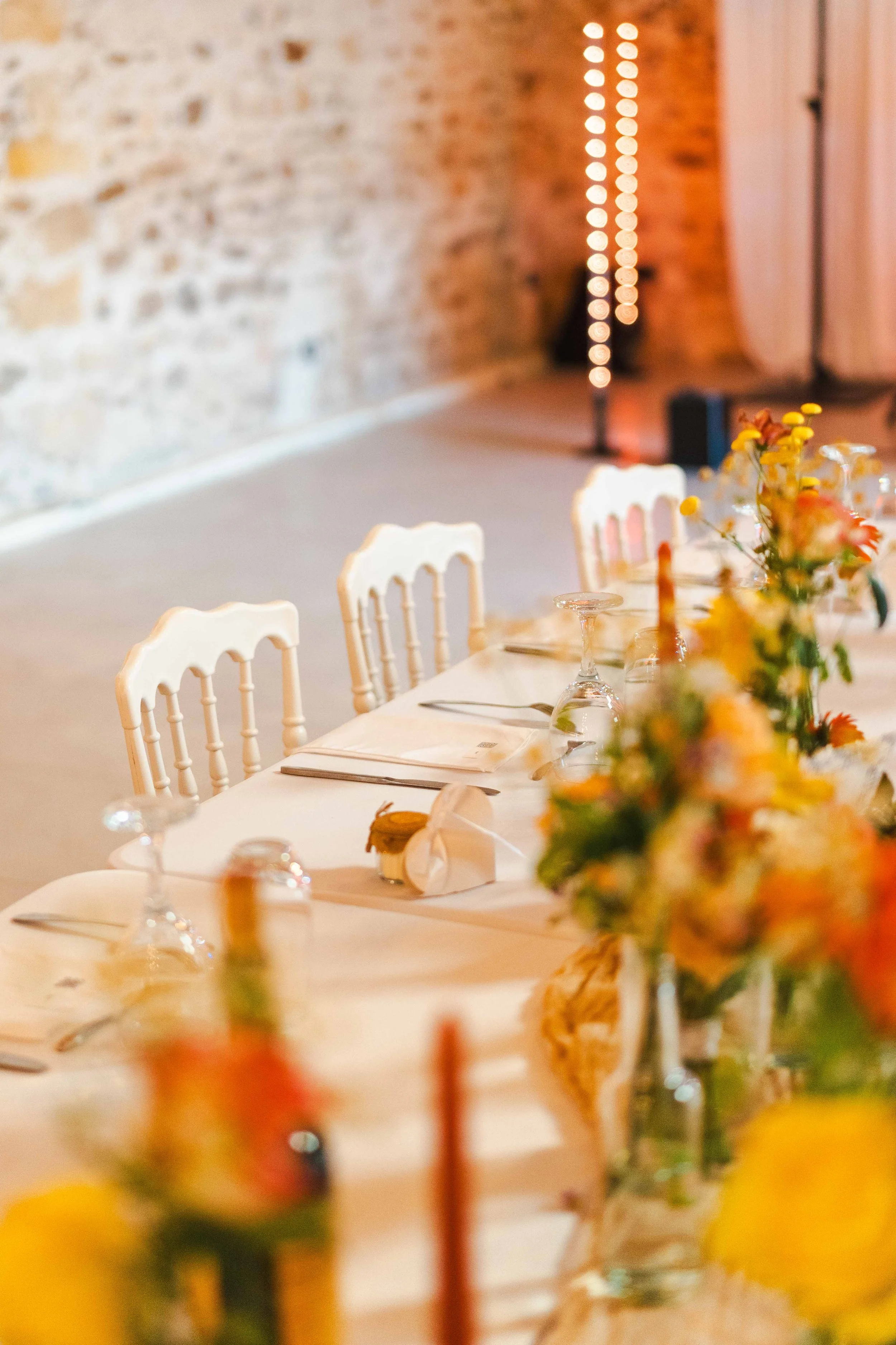 Table dressée avec des assiettes, des verres à vin, des couverts et un centre de table fleuri dans une salle avec mur en pierre et ambiance chaleureuse.