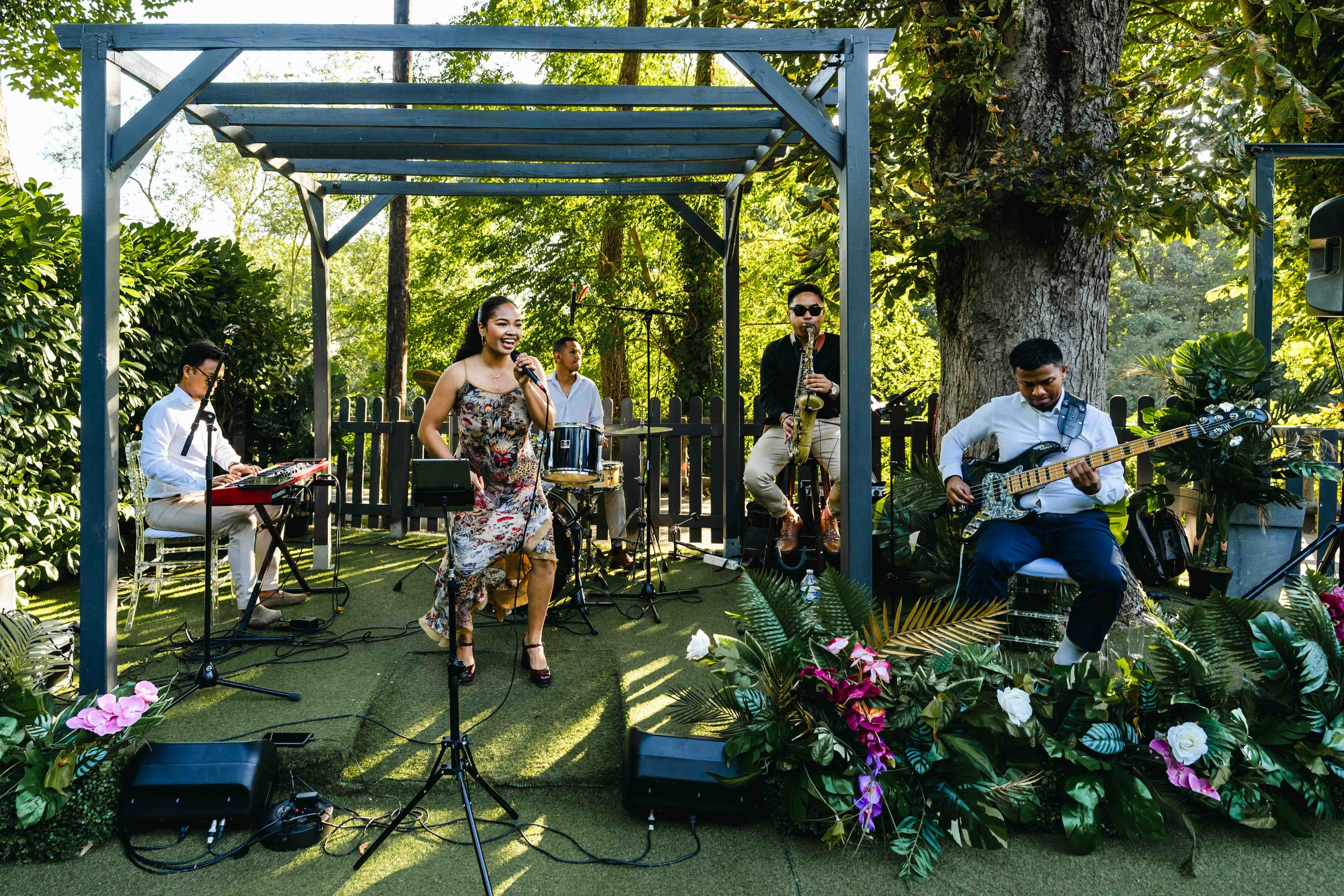 Un groupe de musiciens jouant lors d'une performance en plein air, sous un arbre, entourés de végétation luxuriante et de fleurs au Moulin vert à Champigny.