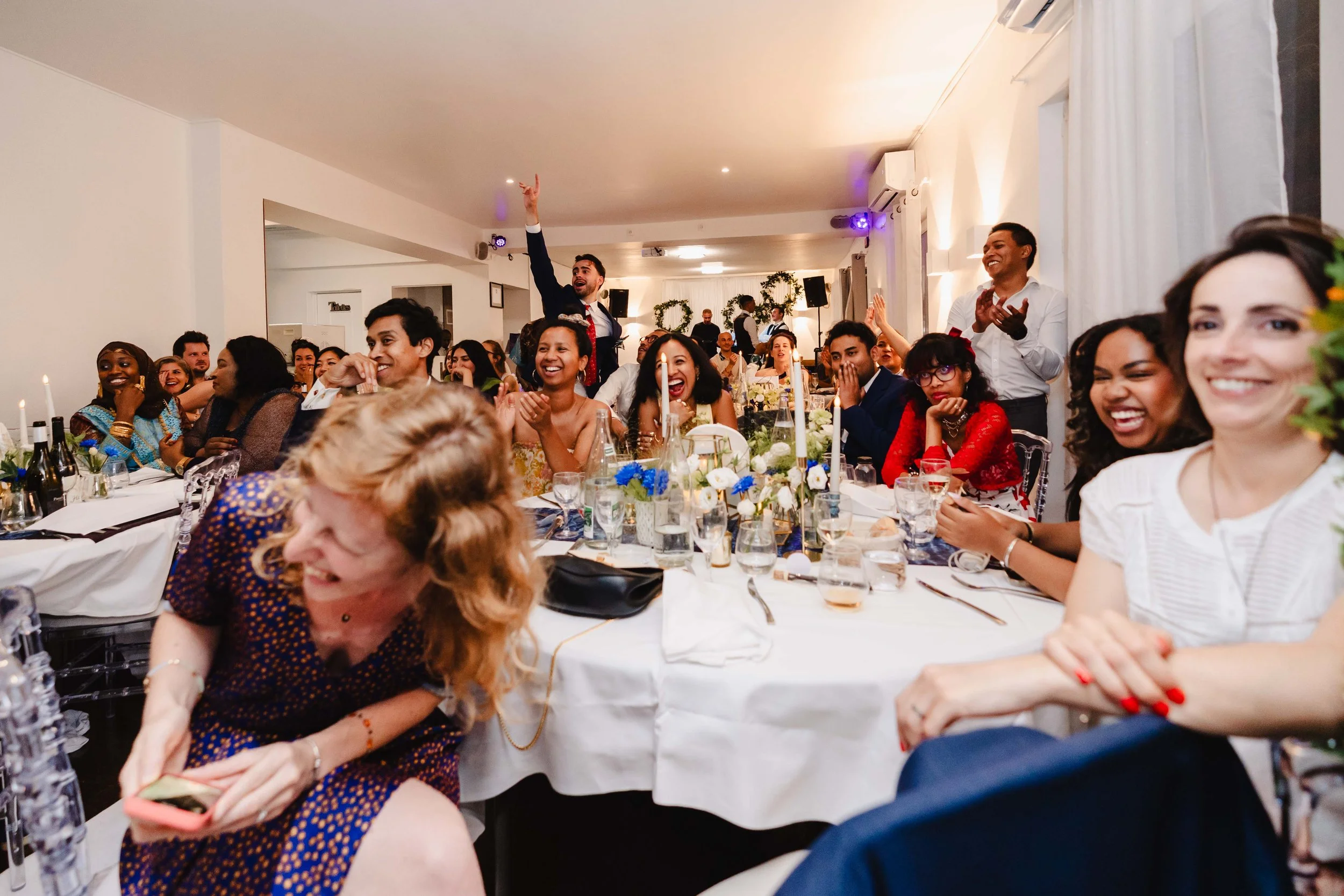 Groupe de personnes assises à une table, riant et applaudissant lors d'une célébration ou d'un mariage, avec un homme debout en arrière-plan faisant un geste joyeux.