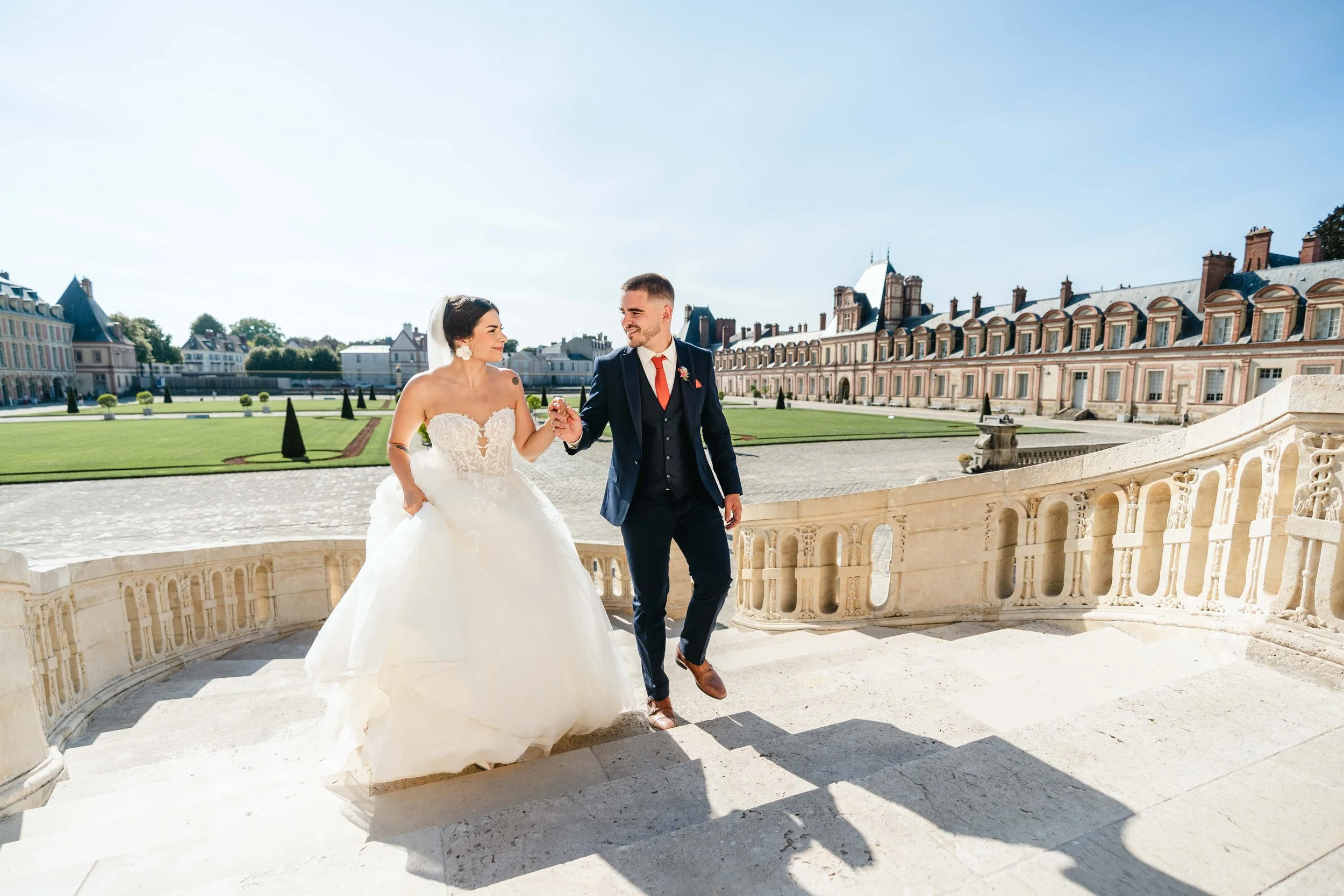 Un couple de mariés entre en tenue de mariage sur l'escalier en pierre du château de Fontainebleau, avec le jardin en arrière-plan, sous un ciel ensoleillé.