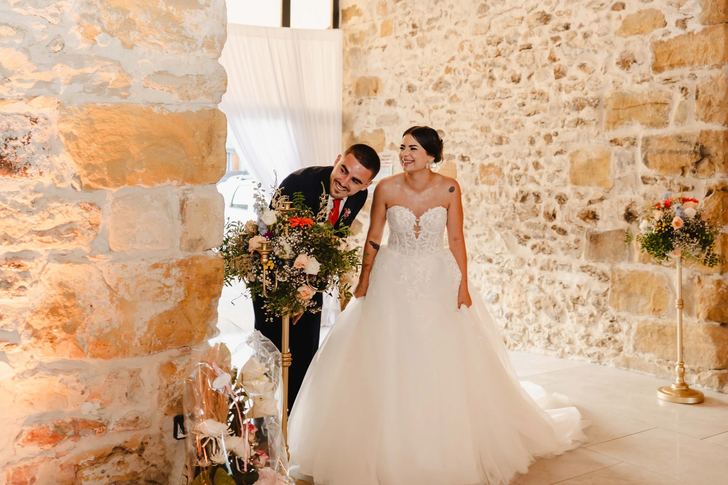 Un couple de mariés souriant à l'intérieur d'une salle avec murs en pierre, décoration florale.