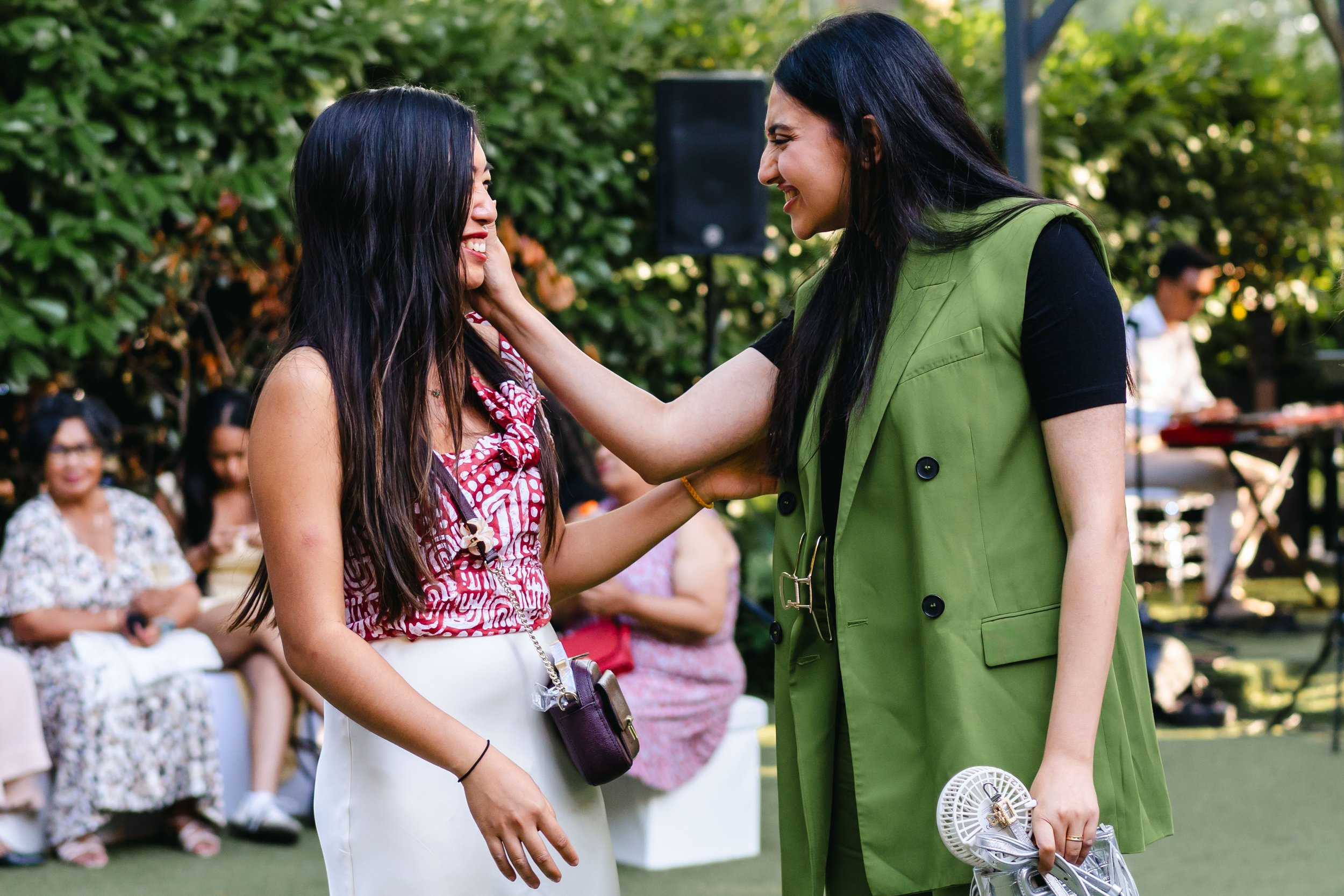 Deux femmes souriant et se touchant la joue lors d'un rassemblement en plein air avec des personnes assises en arrière-plan et de la verdure.