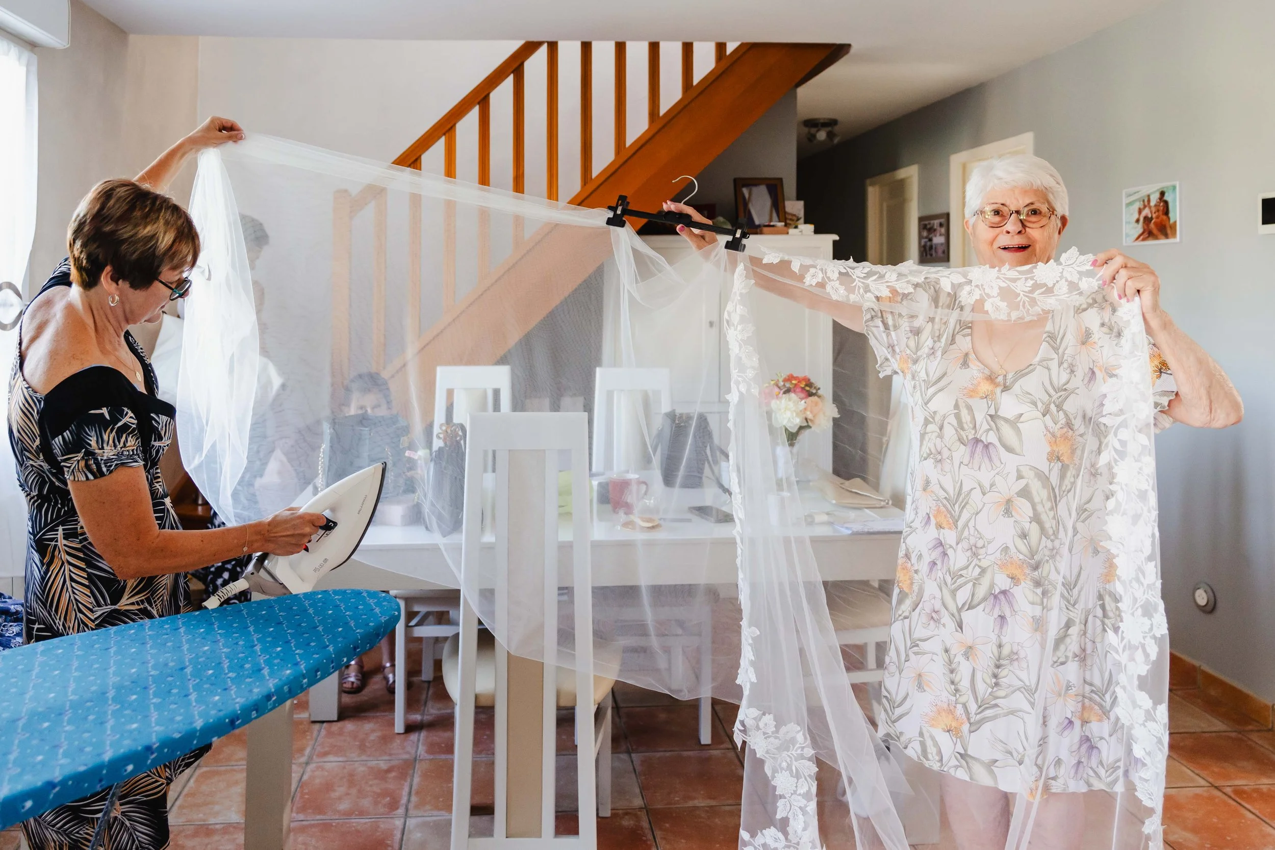 Une personne âgée souriante tient un tissu blanc brodé, probablement une robe de mariée, devant un mur gris dans une maison, tandis qu'une autre personne à gauche plie un vêtement avec un fer à repasser. La scène semble être une préparation pour une 