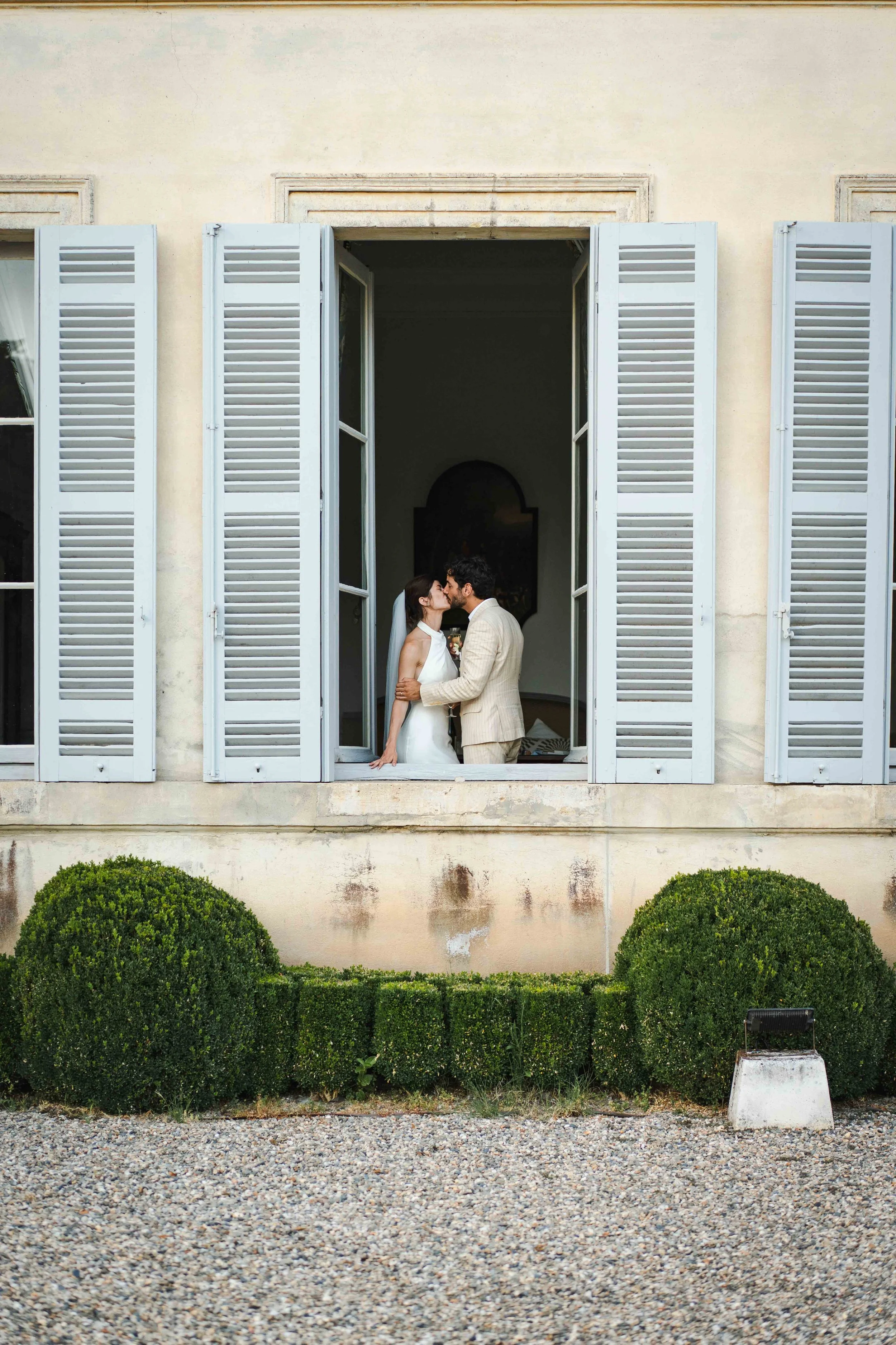 Un couple de mariés partage un baiser dans une grande fenêtre ouverte du chateau Saint Denis, extérieur d'une maison avec des volets en bois gris et des buissons verts.