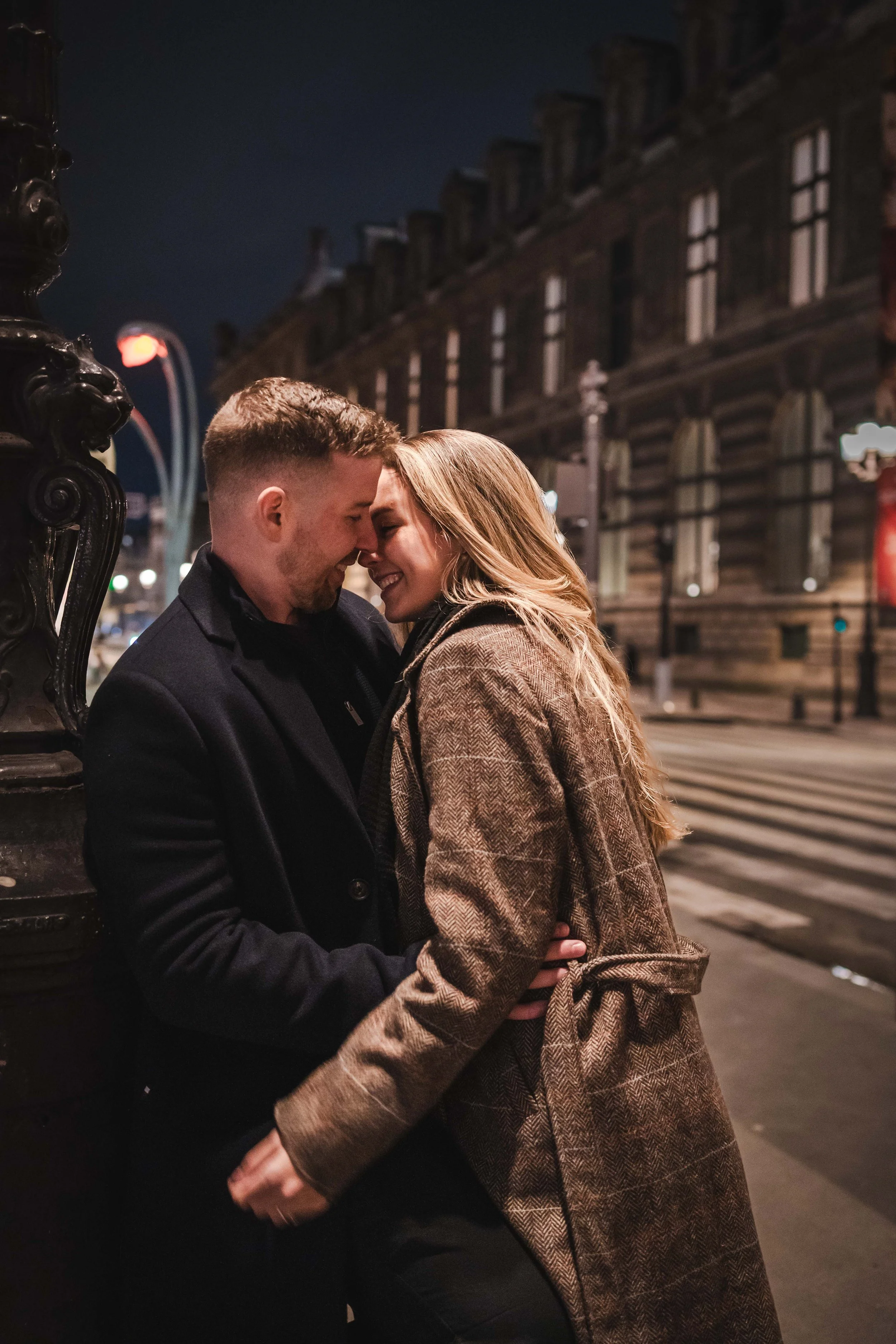 Un couple souriant très proche, se touchant le front, dans une rue à Paris la nuit.