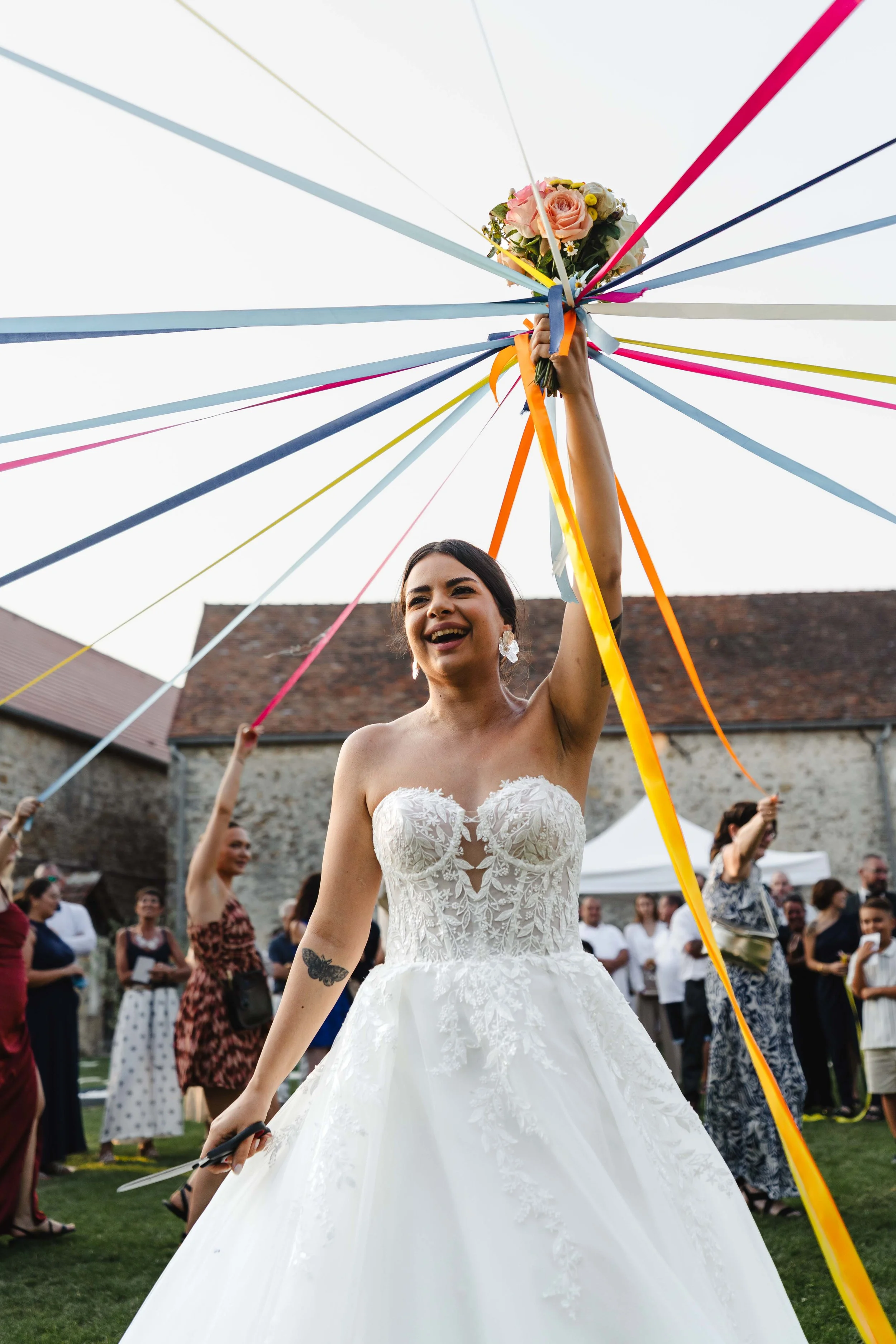 Une femme en robe de mariée souriante, tenant un bouquet de fleurs, sous un cercle de rubans colorés lors d'une célébration extérieure.