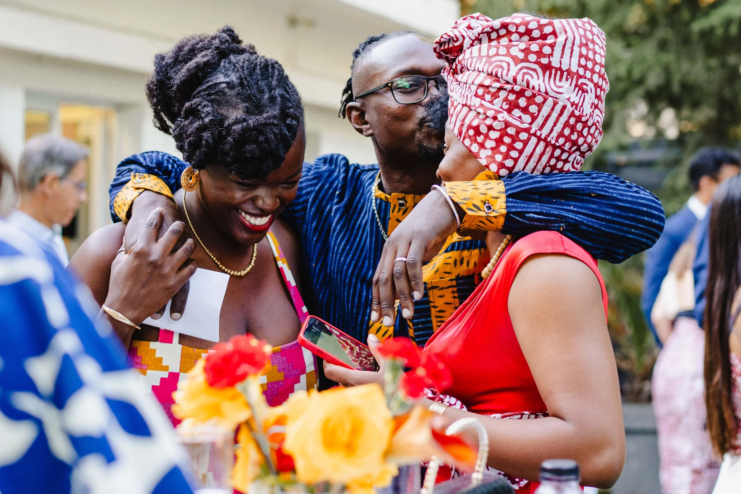Trois personnes souriantes qui s'embrassent et se donnent une poignée de main lors d'une réunion ou célébration en extérieur, avec d'autres personnes en arrière-plan.