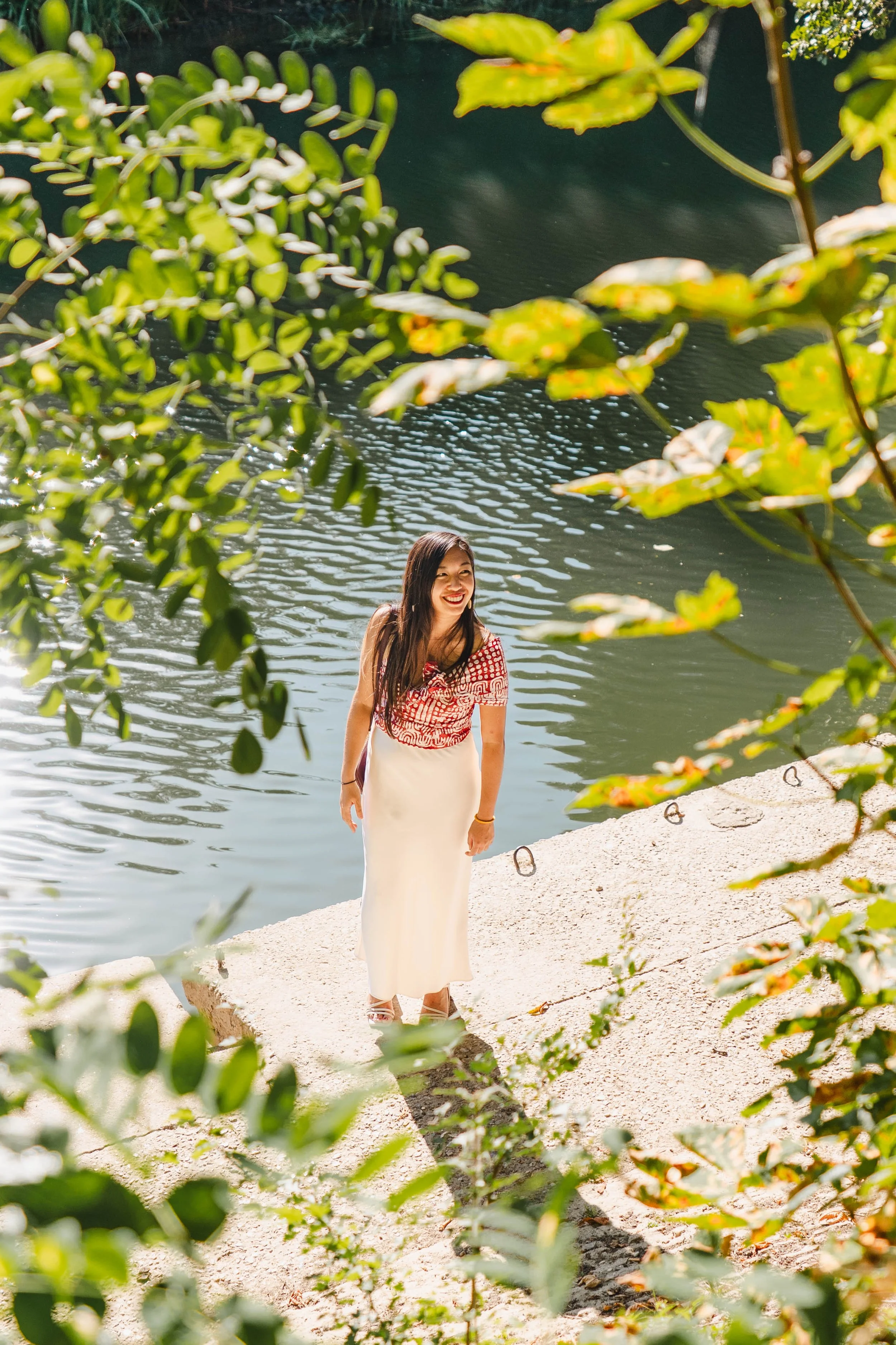 Femme souriante en robe longue blanche, debout sur un rivage rocailleux près de la Marne, à côté de la salle de réception du Moulin vert à Champigny, entourée de feuillage vert et lumineux.