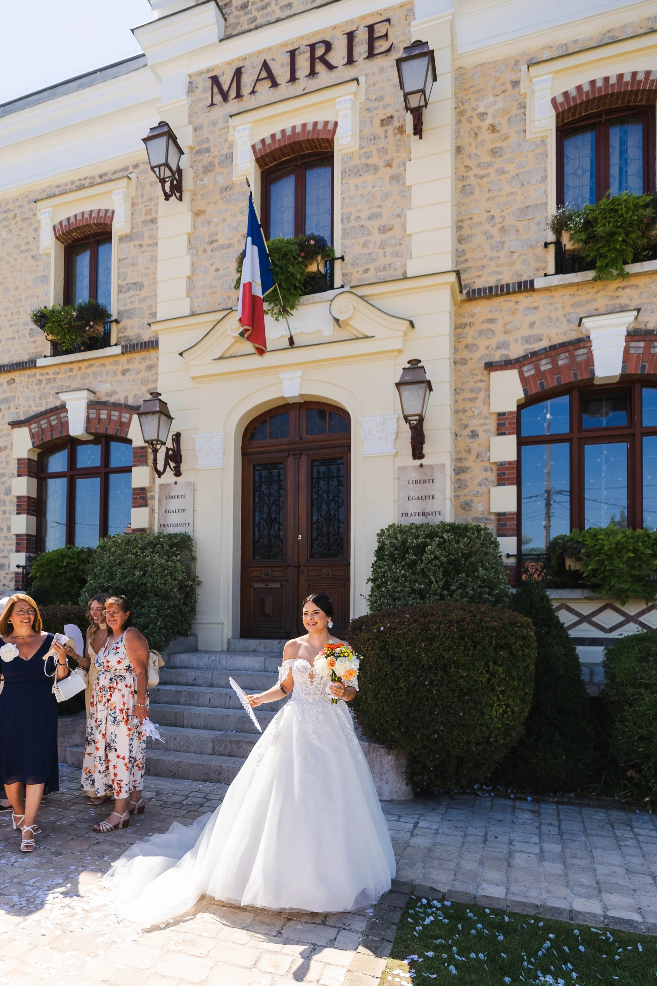 Une mariée en robe blanche tenant un bouquet de fleurs devant la mairie de Bagneux Sur Loing. avec des personnes souriantes autour.