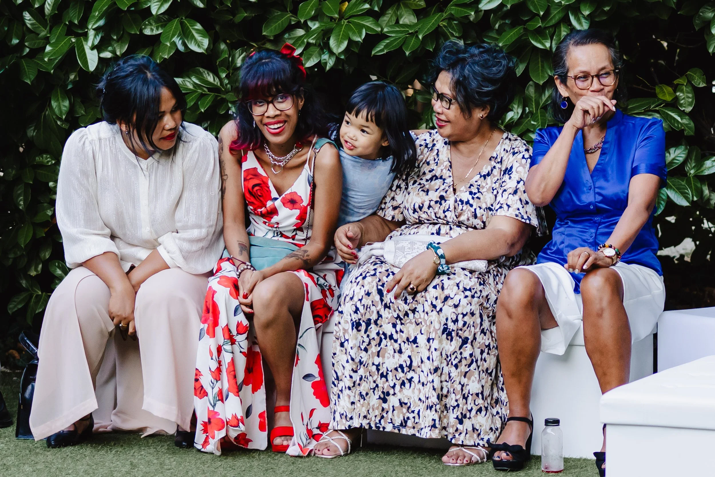 Cinq femmes et un enfant assis sur un banc, entouré de feuillage vert, partageant un moment joyeux.