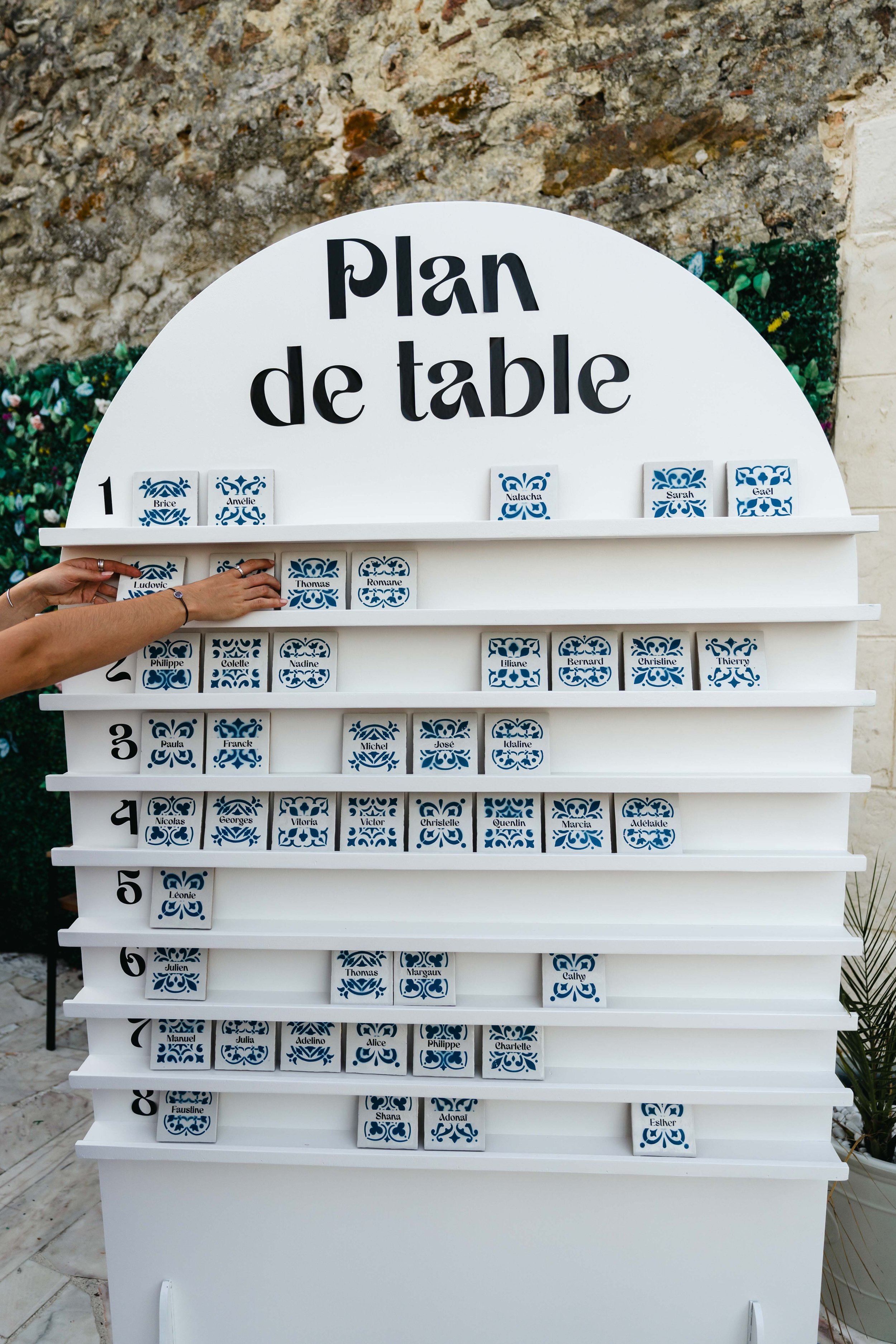 Plan de table de mariage avec noms en céramique bleue sur un tableau blanc, disposé par numéro de table et nom.